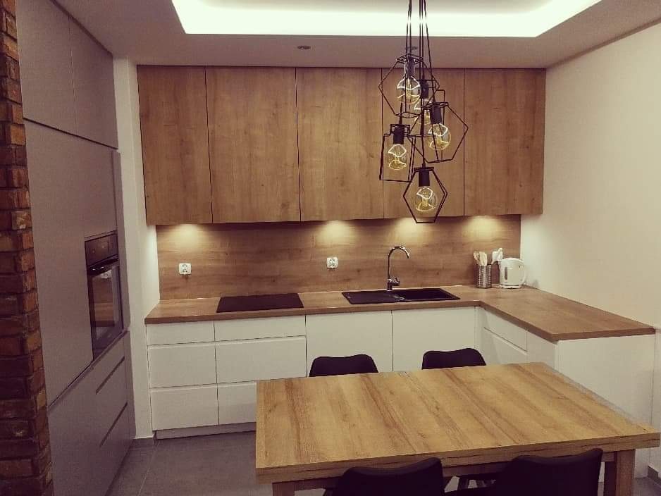 Nowoczesna kuchnia z drewnianymi szafkami górnymi i białymi dolnymi, oświetlona lampami wiszącymi w geometrycznych kloszach, z drewnianym stołem i krzesłami w centralnej części.