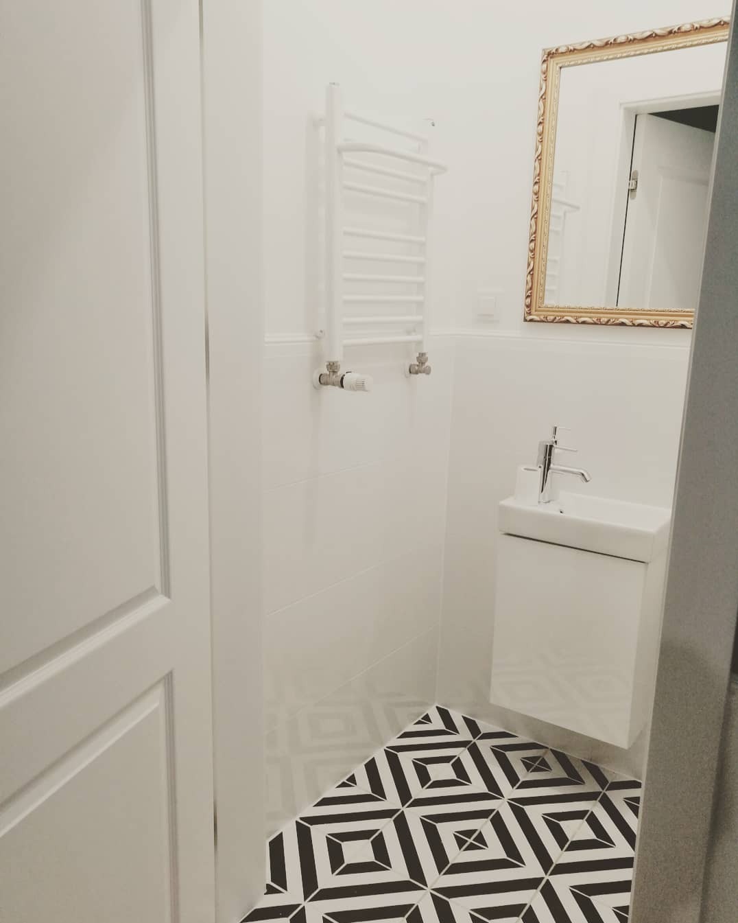Nowoczesna, biała łazienka z geometryczną, czarno-białą podłogą, wiszącą umywalką i grzejnikiem drabinkowym, dopełniona lustrem w złotej ramie.