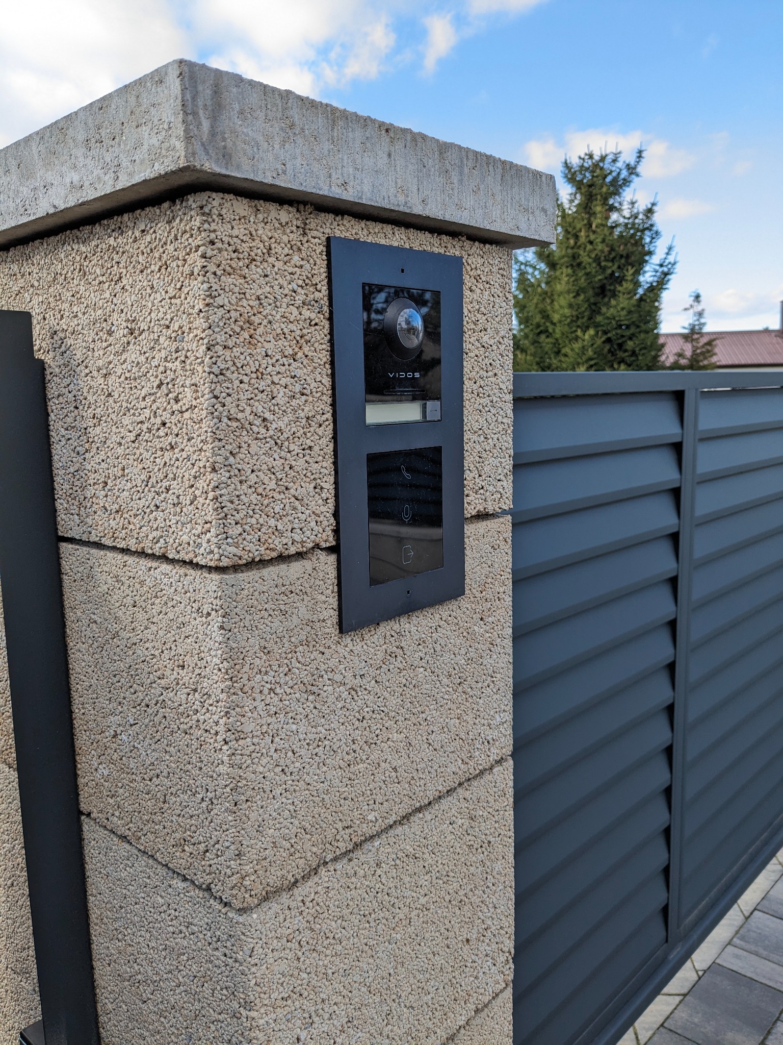 Zewnętrzny panel wideodomofonu Vidos zamontowany na słupku ogrodzeniowym z betonu architektonicznego, obok przesuwna brama lamelowa w kolorze antracytowym.