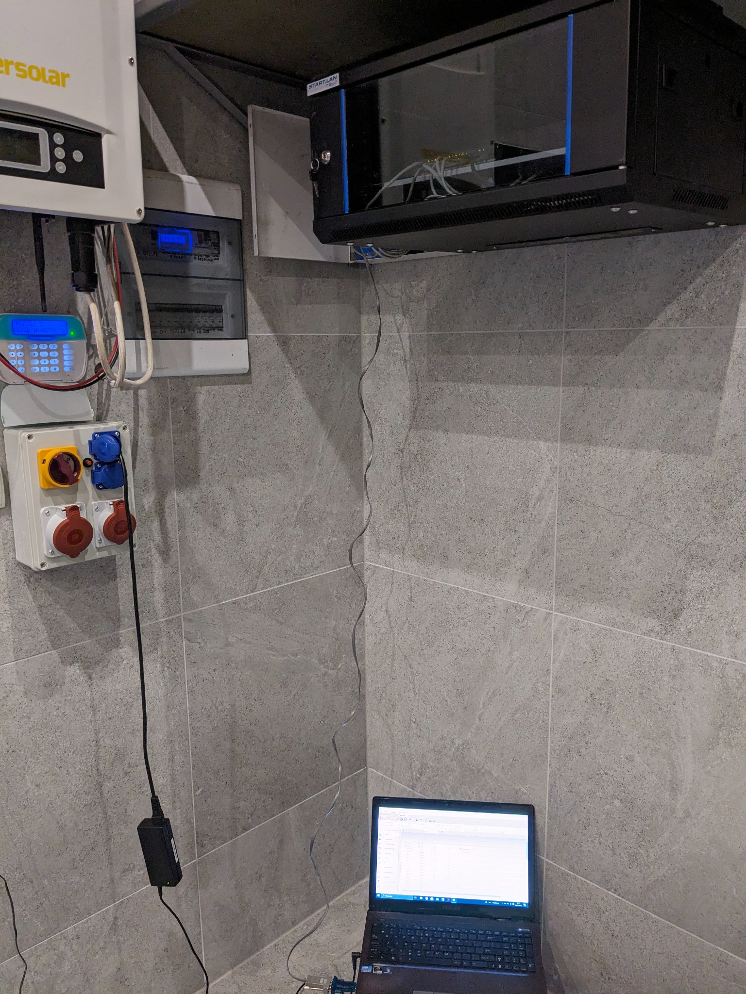 Panel elektryczny z zabezpieczeniami, skrzynka z gniazdami siłowymi i niskonapięciowymi, szafa rack z okablowaniem sieciowym oraz laptop z uruchomionym oprogramowaniem diagnostycznym na tle szarej...