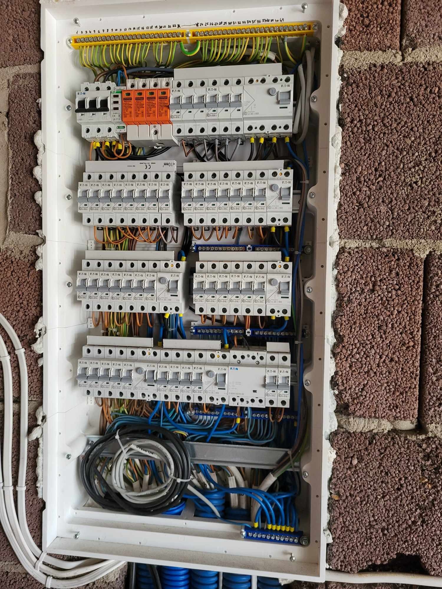 Otwarta skrzynka elektryczna z widocznymi bezpiecznikami, przewodami różnej grubości i koloru, listwami zaciskowymi, zamontowana na ścianie z czerwonej cegły o porowatej strukturze, zabezpieczona...