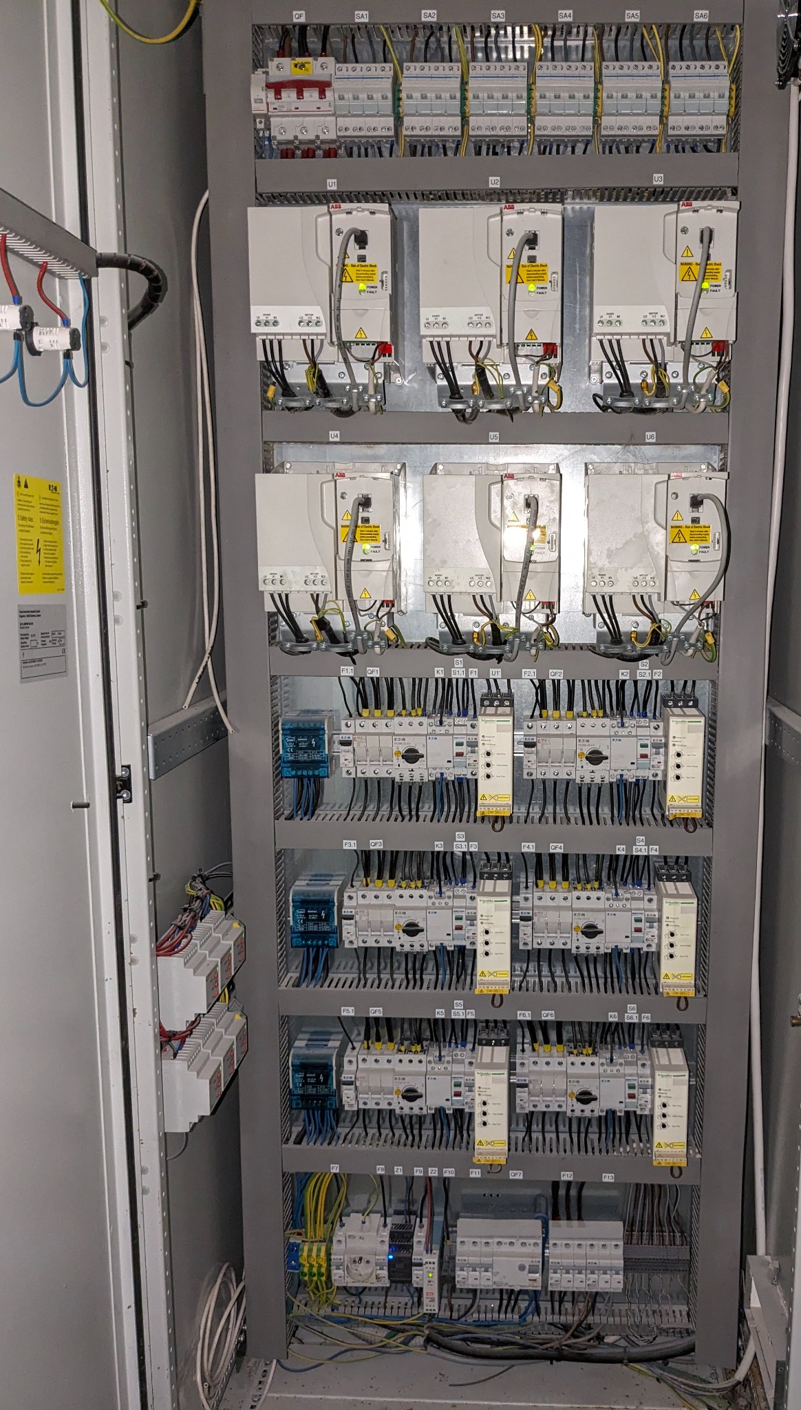 Rozdzielnica elektryczna z widocznymi zabezpieczeniami, modułami ABB i przewodami w kolorach czarnym, żółto-zielonym i niebieskim, zamontowana w szarej szafie metalowej.