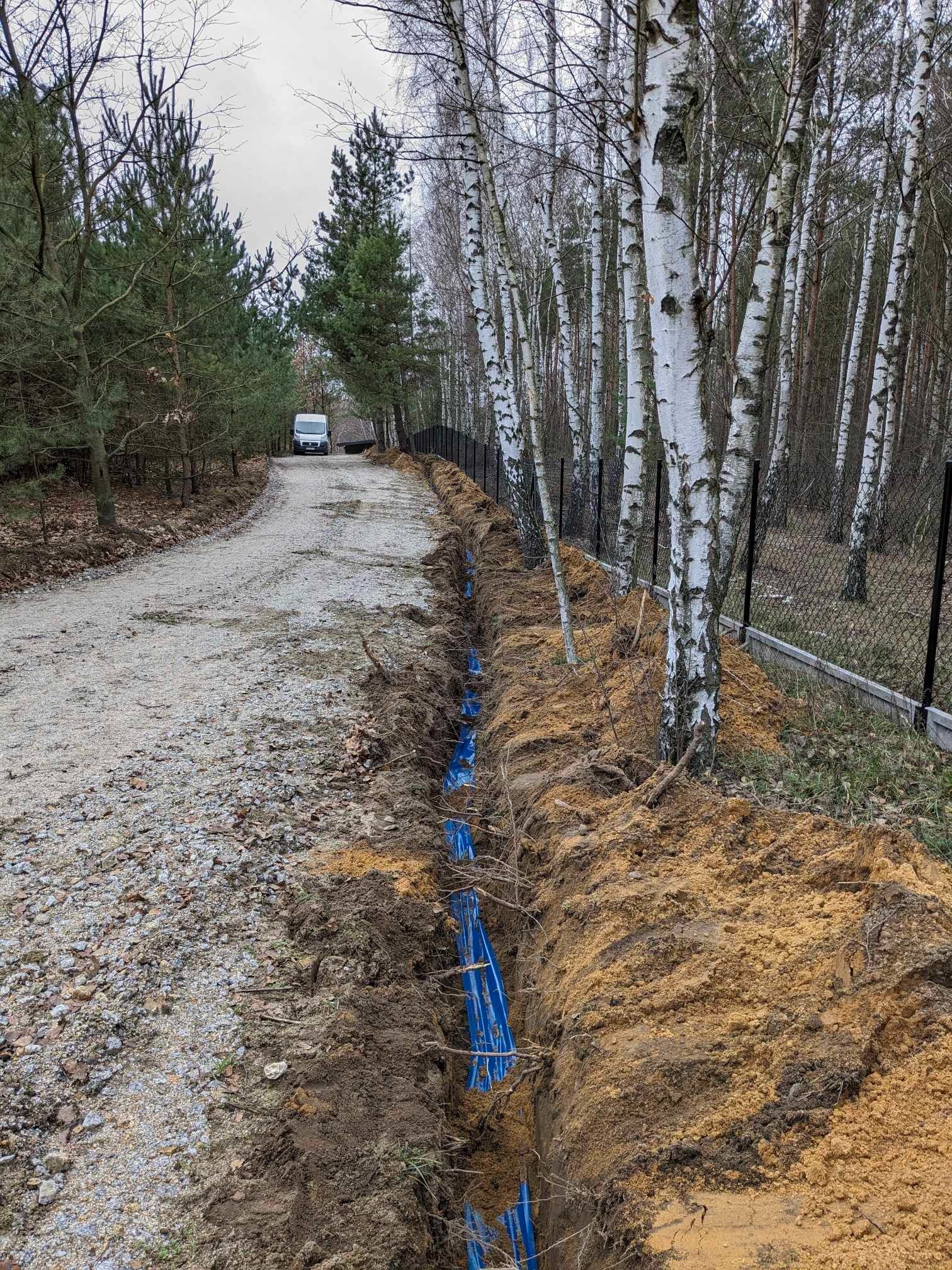 Wykop w ziemi z ułożonymi niebieskimi rurami wzdłuż ogrodzenia z siatki, na tle lasu brzozowego i drogi gruntowej z zaparkowanym busem w oddali.