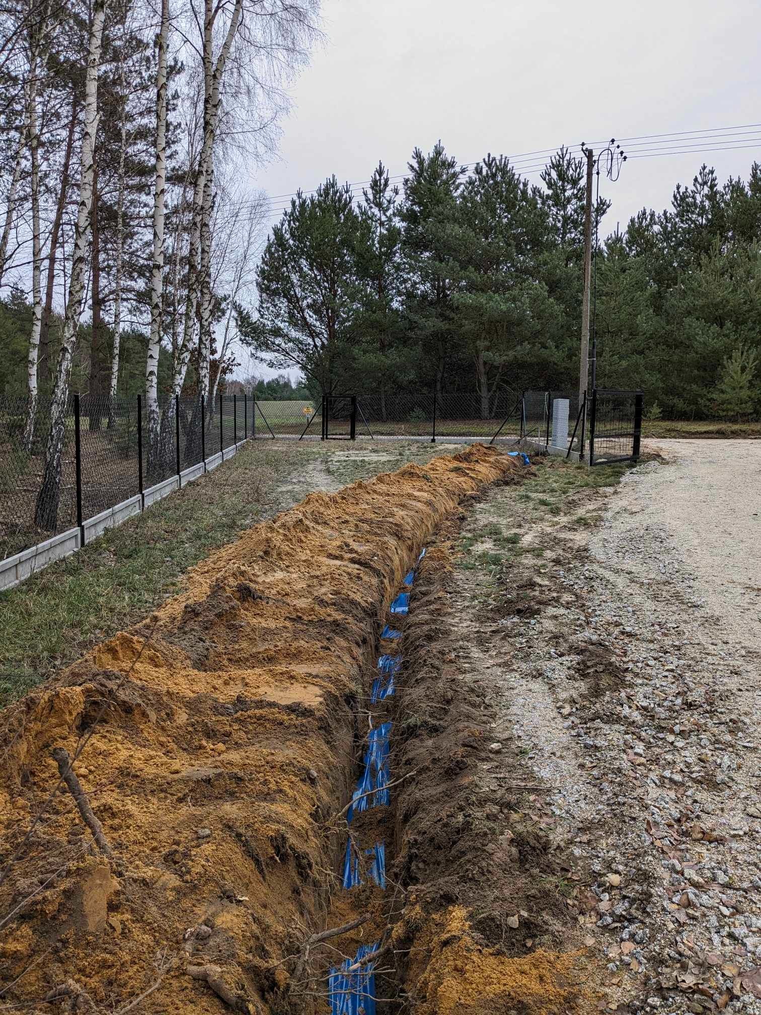 Wykop w ziemi z ułożonymi niebieskimi rurami, prawdopodobnie w trakcie układania przyłącza, widoczny teren działki z ogrodzeniem i drzewami w tle.