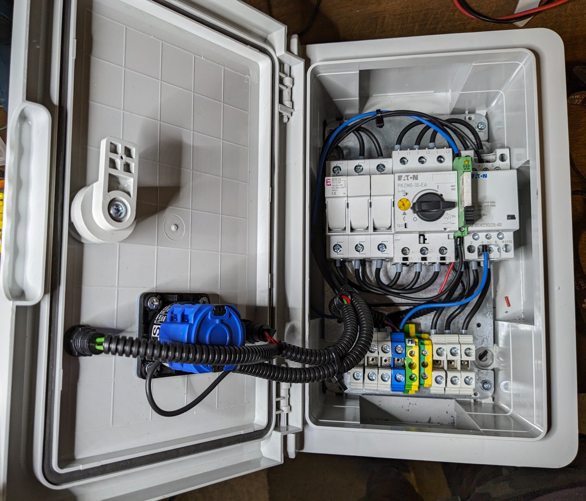 Otwarta skrzynka elektryczna z zamontowanymi bezpiecznikami, wyłącznikiem i listwami zaciskowymi, widoczne kable w peszlach i przewody podłączone do elementów, niebieski element zabezpieczający...