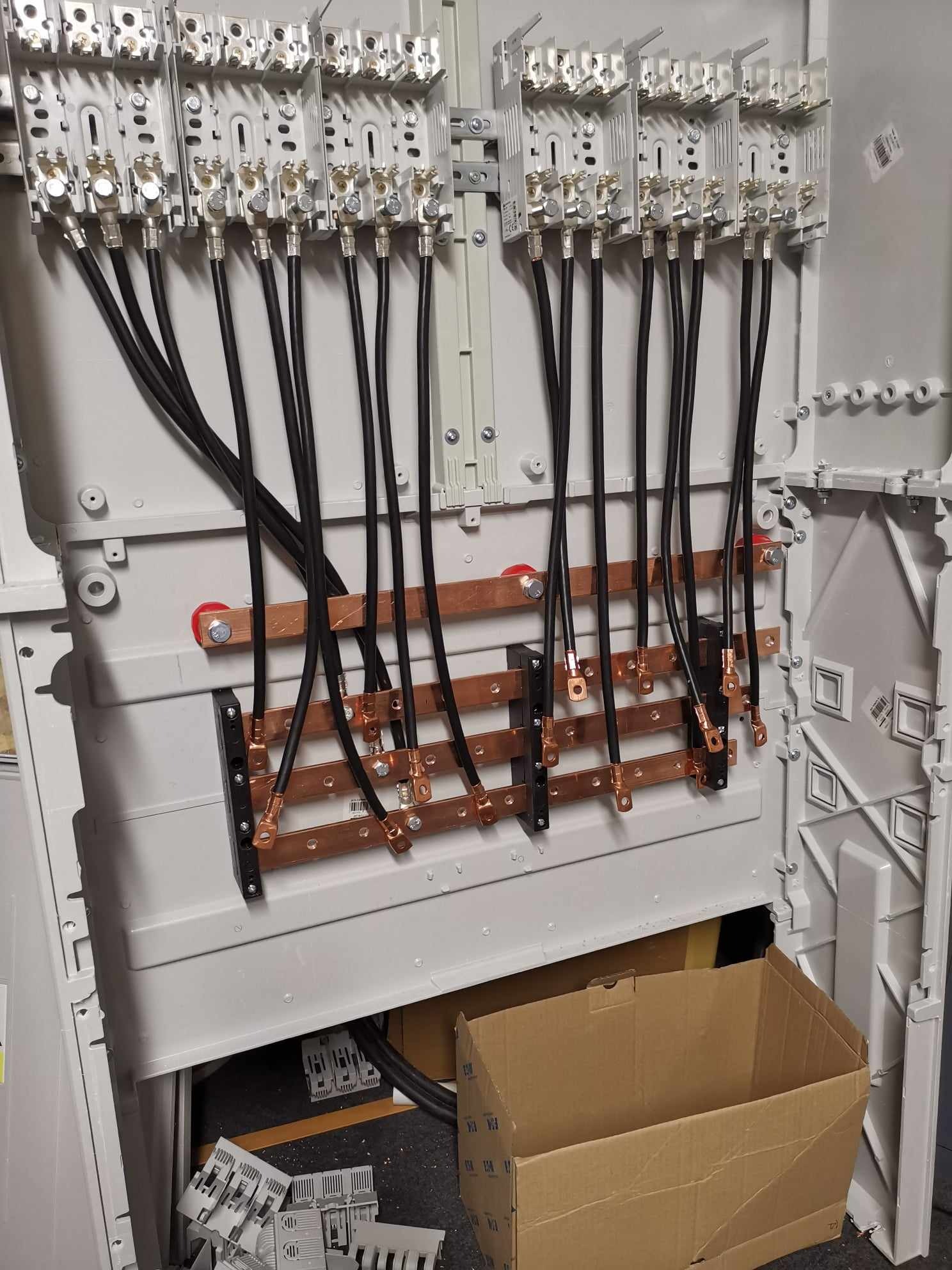 Wnętrze rozdzielnicy elektrycznej z zamontowanymi szynami zbiorczymi miedzianymi i podłączonymi przewodami w czarnej izolacji, widoczna kartonowa skrzynka w dolnej części obudowy.