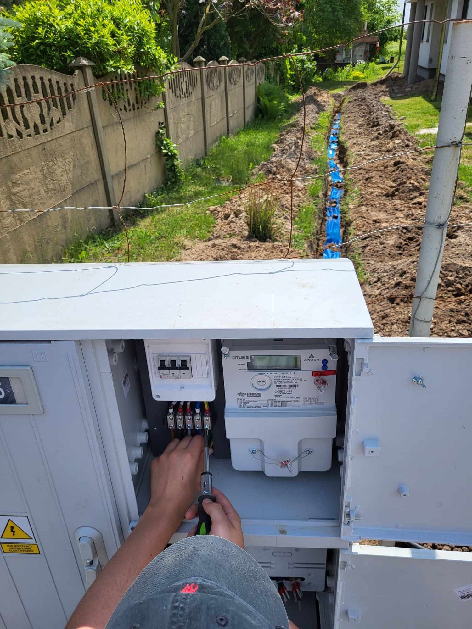 Elektryk podłącza przewody w szafce z licznikiem elektrycznym na tle wykopanego rowu z niebieską rurą kablową.