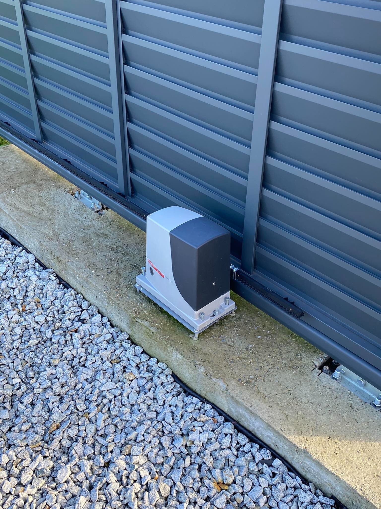 Szary automat do bramy przesuwnej z łańcuchem zębatym, zamontowany na betonowej podstawie obok szarego ogrodzenia panelowego, widok z bliska na tle żwiru.