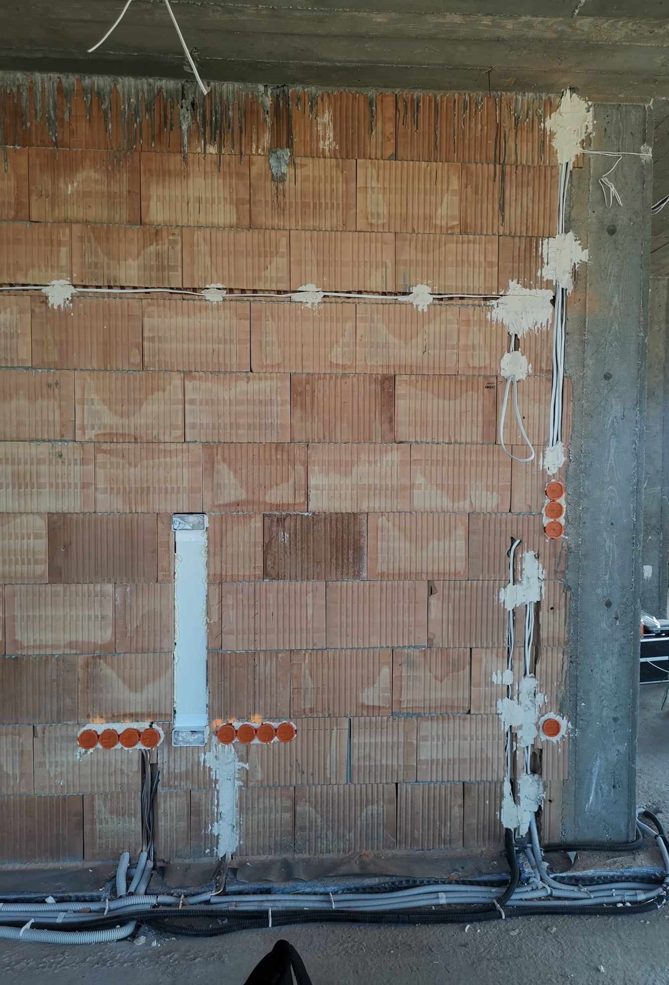 Murowana ściana w trakcie prac elektrycznych: widoczne kable w peszlach, puszki instalacyjne zalane gipsem, pionowy kanał kablowy w ścianie.