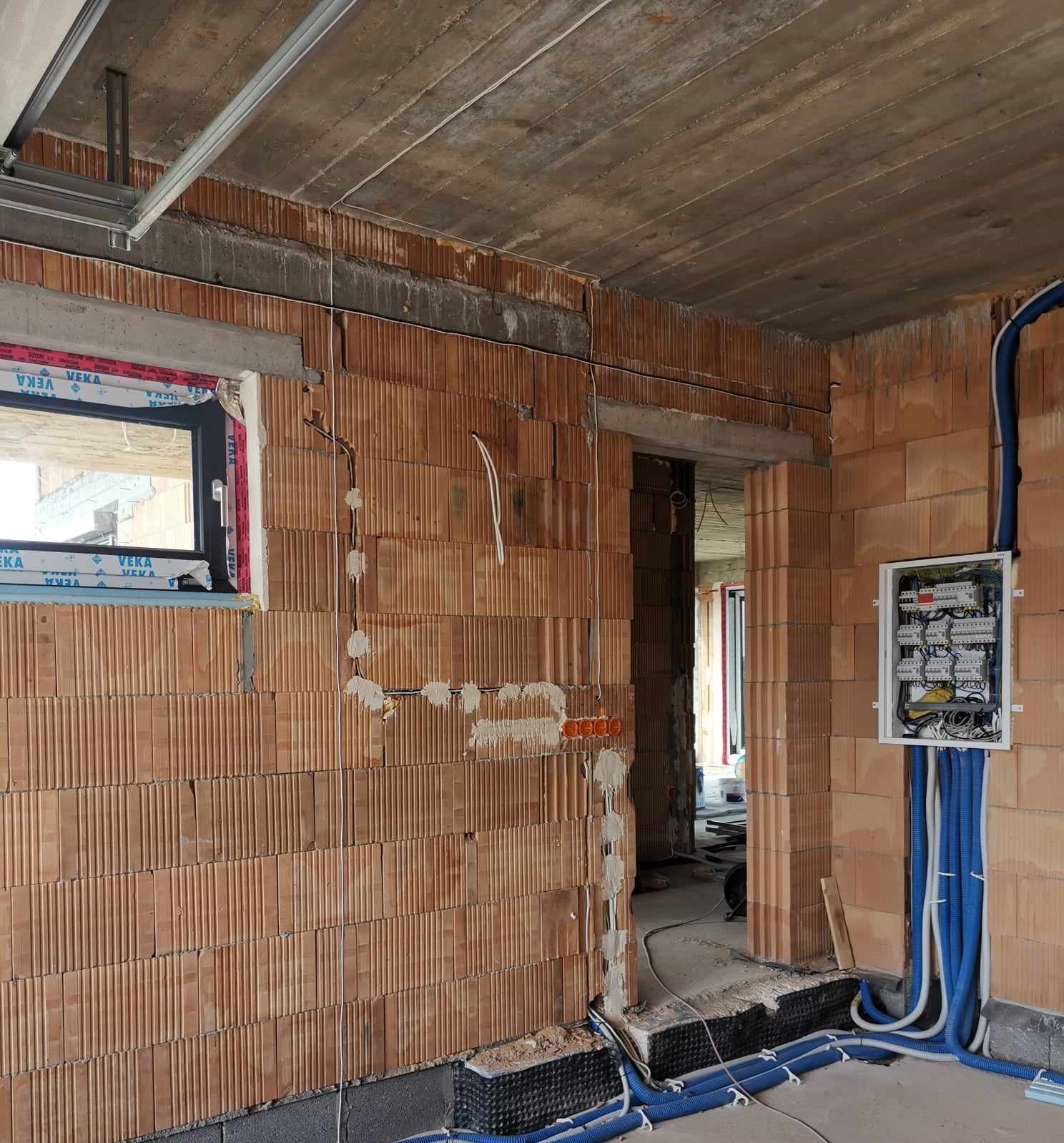 Wnętrze w trakcie budowy: ściany z cegły, okno z folią ochronną VEKA, instalacja elektryczna w rurach karbowanych, rozdzielnica elektryczna na ścianie, widoczne puszki instalacyjne.