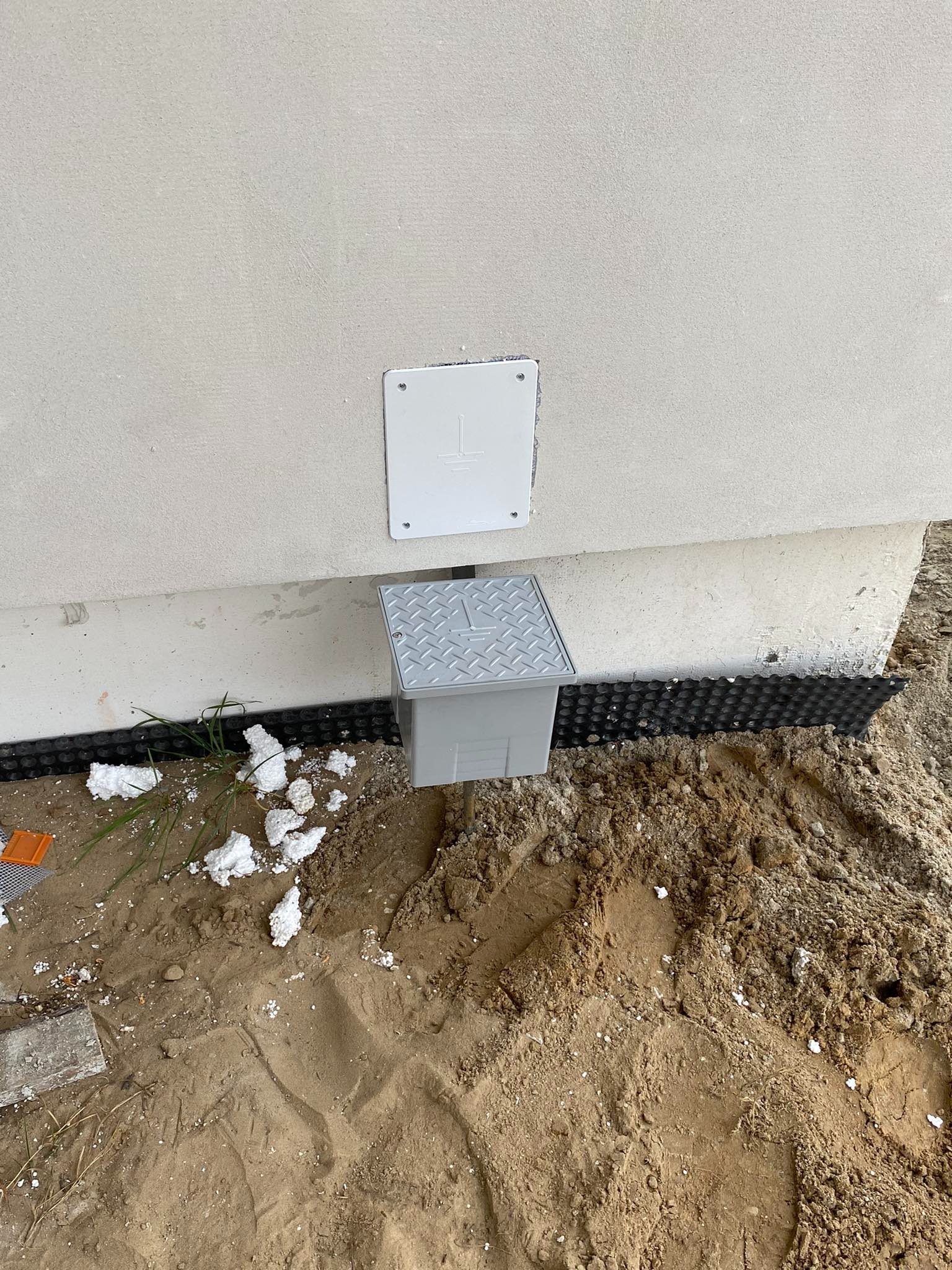 Szara skrzynka z pokrywą o wzorze rombów, wystająca z ziemi przy ścianie budynku, widoczna czarna folia kubełkowa, biała puszka elektryczna z symbolem uziemienia na ścianie, fragmenty styropianu...