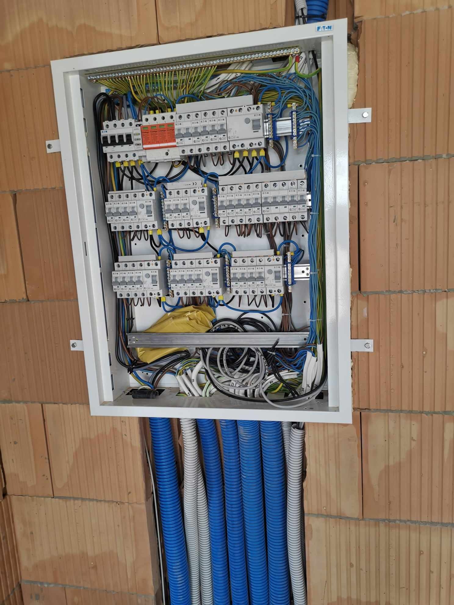 Otwarta skrzynka rozdzielcza z licznymi bezpiecznikami, przewodami i niebiesko-szarymi rurami karbowanymi, zamontowana na ścianie z cegły kratówki.