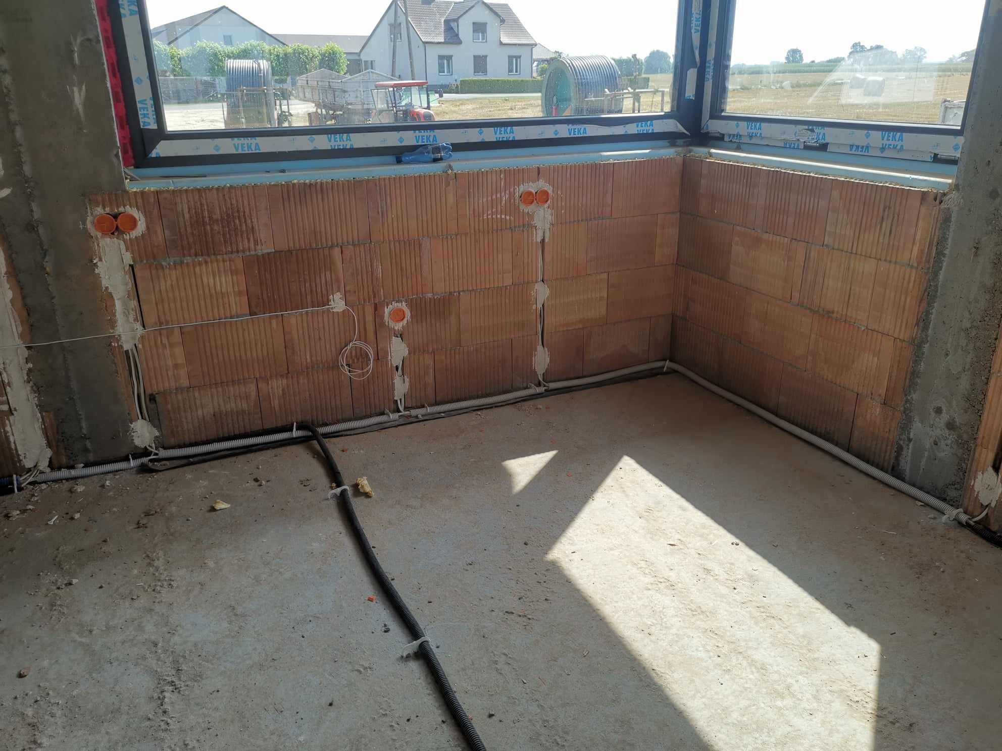 Wewnętrzne ściany budynku w trakcie instalacji elektrycznej, widoczne pomarańczowe puszki instalacyjne i peszle prowadzące kable, surowy beton na podłodze i okno z widokiem na wiejski krajobraz.