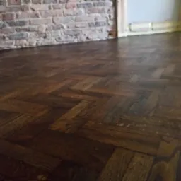 Parkiet ułożony w jodełkę po cyklinowaniu, z widoczną starą ceglaną ścianą w tle i zabezpieczonymi taśmą malarską framugami drzwi.