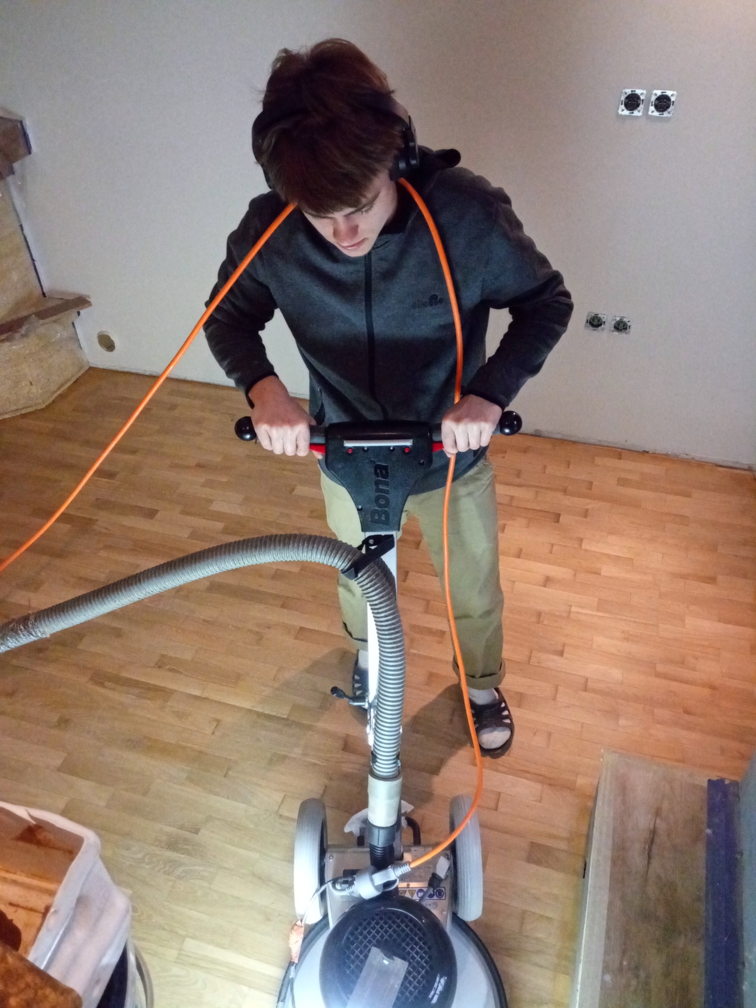 Młody mężczyzna w słuchawkach cyklinuje parkiet maszyną Bona, widoczne gniazdka elektryczne na ścianie