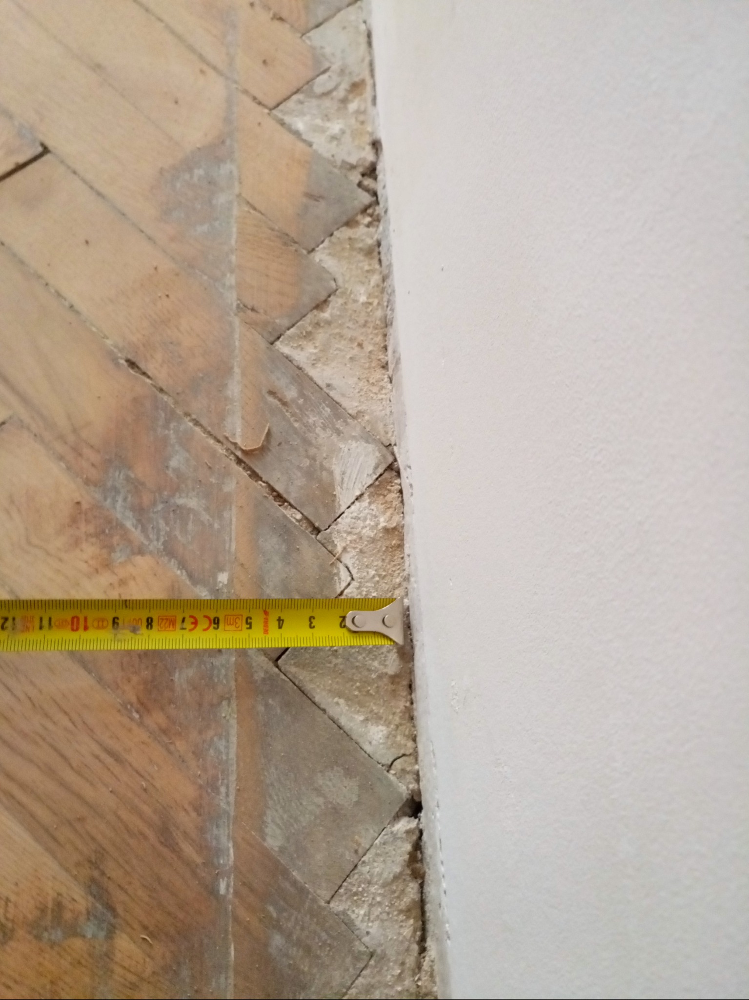 Pomiar odległości od ściany do parkietu ułożonego w jodełkę, widoczny fragment ściany i żółta miarka zwinięta do 2 cm.