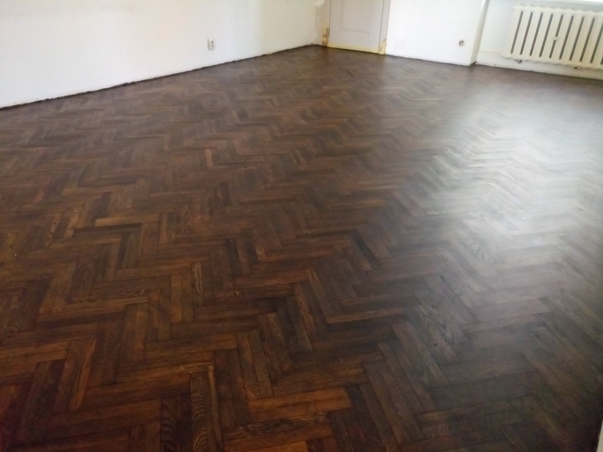 Odnowiona, ciemnobrązowa podłoga parkietowa ułożona we wzór jodełki, widoczna w pustym pokoju z białymi ścianami i grzejnikiem.
