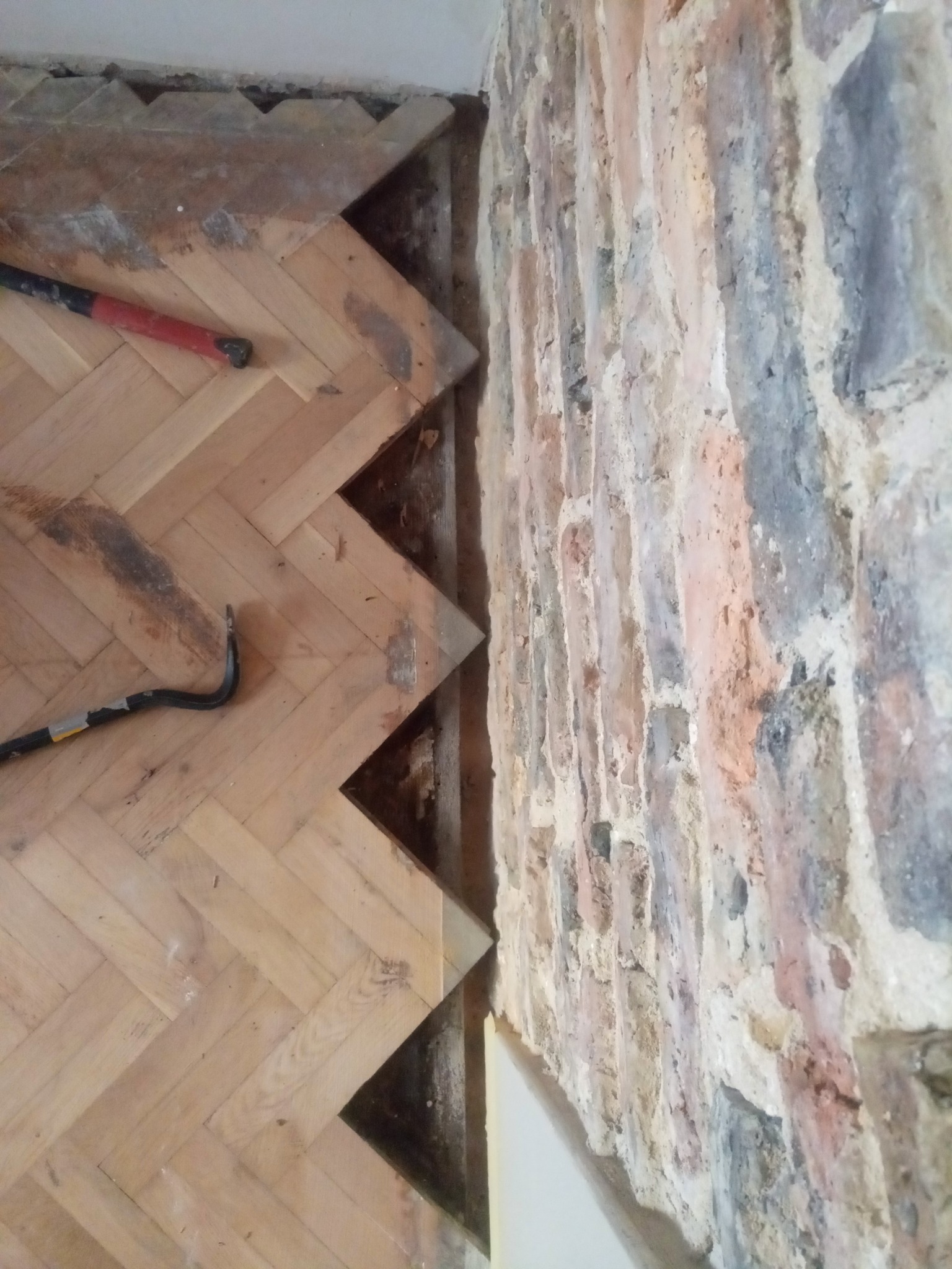 Fragment podłogi z parkietu ułożonego w jodełkę, z widocznymi śladami po usuniętych listwach przypodłogowych i narzędziami: młotkiem i łomem. Obok odsłonięta, surowa ściana z cegły o zróżnicowanej...