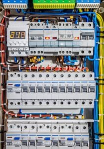 Rozdzielnia elektryczna z licznymi bezpiecznikami, przewodami w różnych kolorach i wyświetlaczem cyfrowym wskazującym wartość. Układ schludny i uporządkowany, widoczne oznaczenia na elementach.