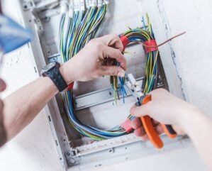 Elektryk w niebieskiej czapce łączy kolorowe przewody w skrzynce elektrycznej za pomocą pomarańczowych szczypiec, widoczne zegarek na ręku i biała ściana w tle.