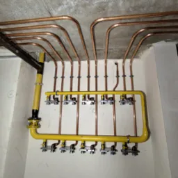 Miedziane rury instalacji grzewczej pod stropem, z żółtą magistralą i zaworami. Widok na surowy betonowy sufit i fragment ściany.