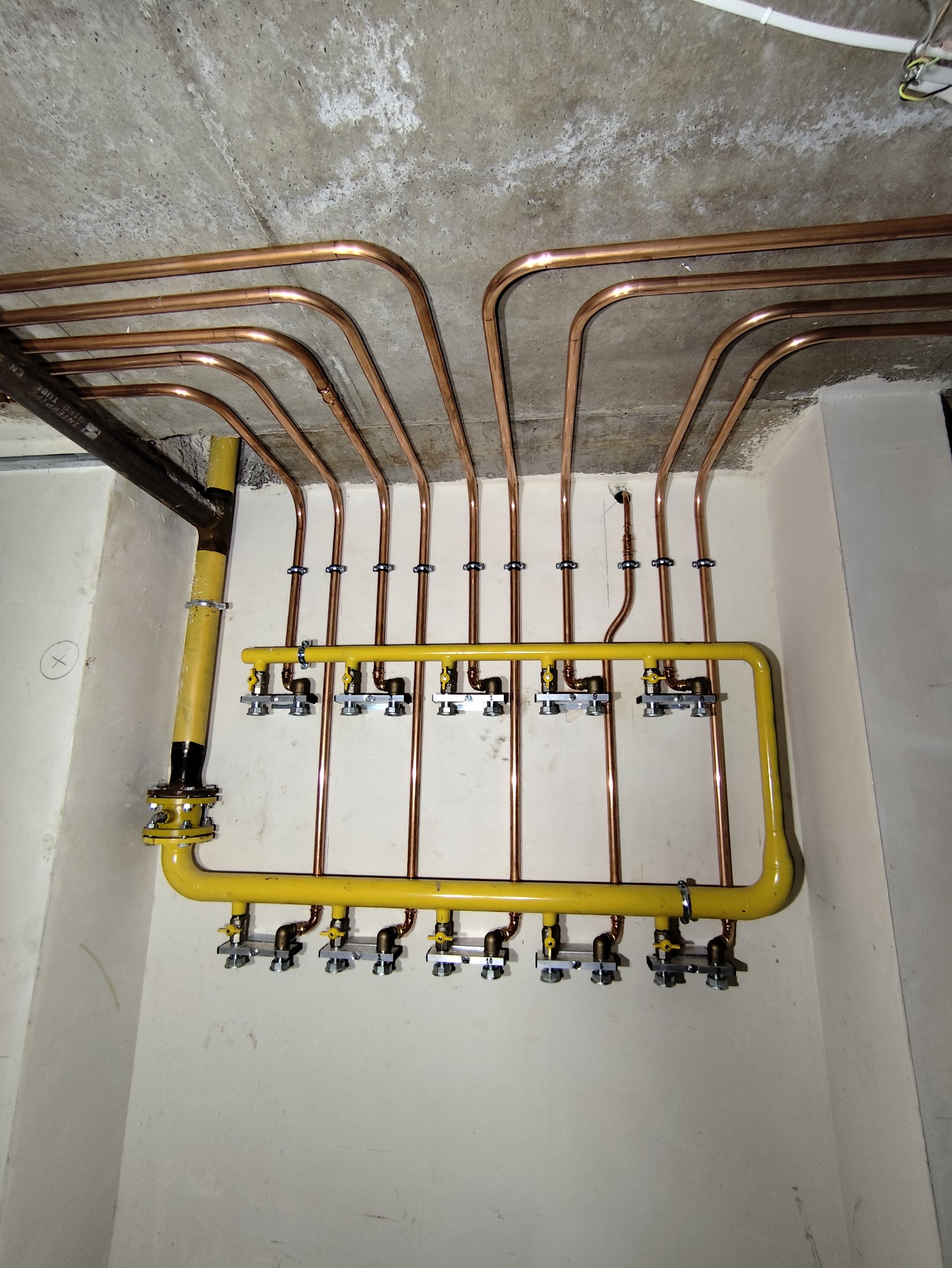 Miedziane rury instalacji grzewczej pod stropem, z żółtą magistralą i zaworami. Widok na surowy betonowy sufit i fragment ściany.