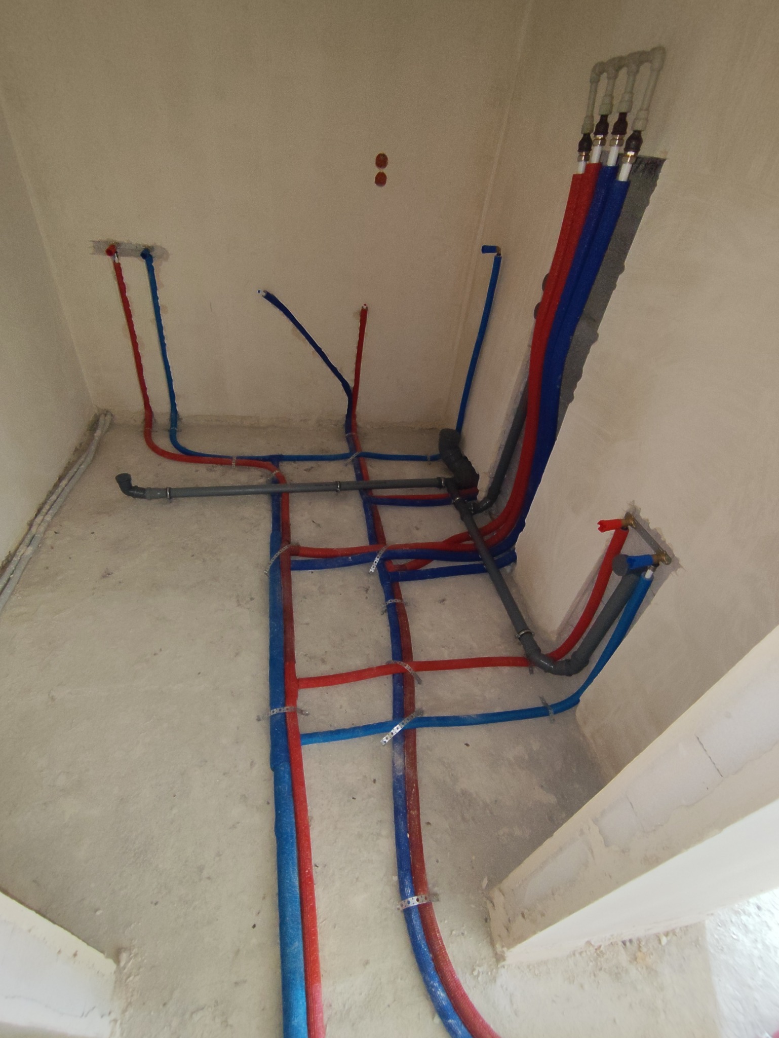 Rozbudowana instalacja wodna w surowym wnętrzu: liczne rury (czerwone i niebieskie) prowadzone po podłodze i ścianach, częściowo zamocowane metalowymi uchwytami.