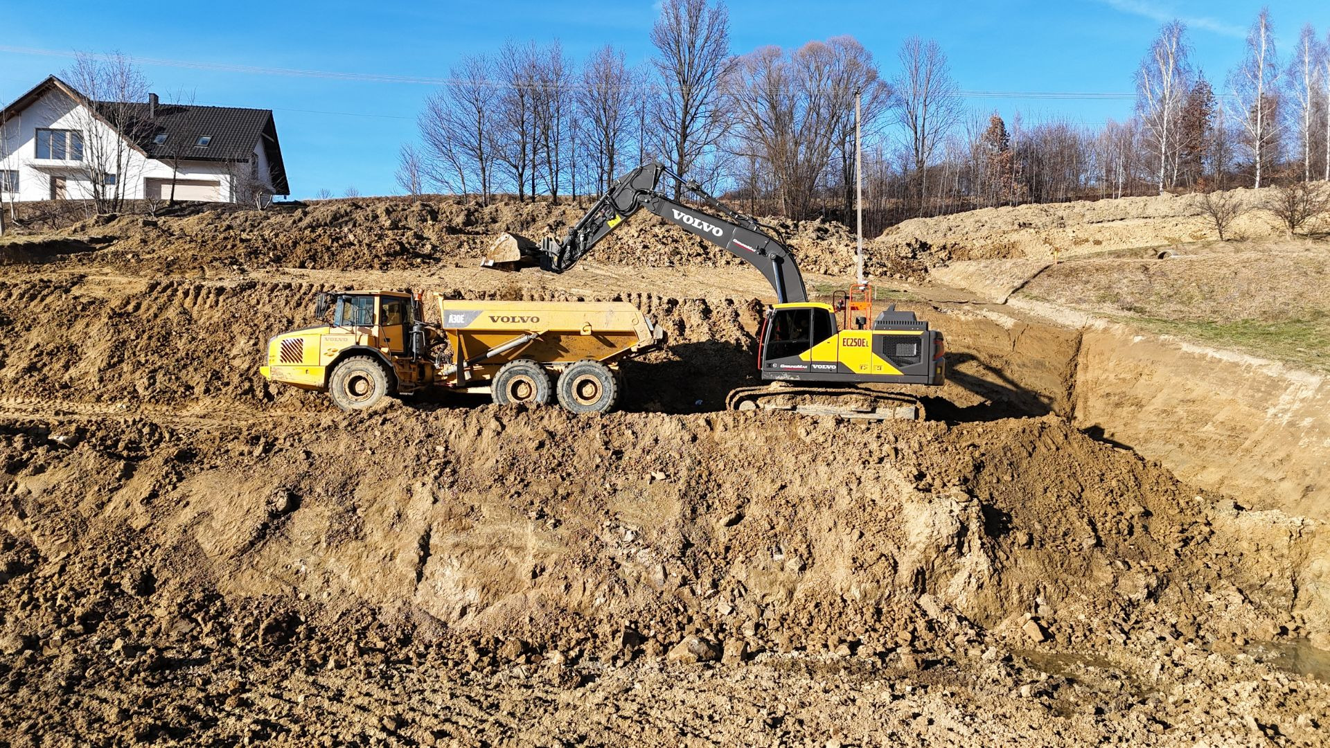 Żółta wywrotka Volvo A30E i koparka Volvo EC250E pracują przy wykopie ziemnym w słoneczny dzień, w tle widoczny dom i drzewa bez liści.
