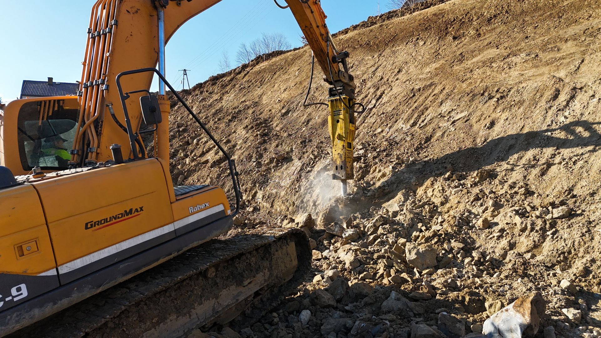 Żółta koparka Hyundai Robex z młotem hydraulicznym kruszy skalisty grunt na zboczu, w tle widoczny fragment budynku.