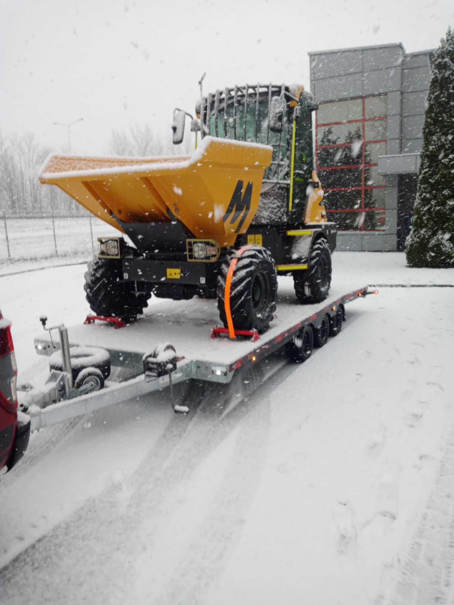 Żółty wozidło budowlane zabezpieczone pasami transportowymi na niskopodwoziowej przyczepie w Krzeczowie podczas śnieżnej pogody, na drugim planie budynek z szarą elewacją.
