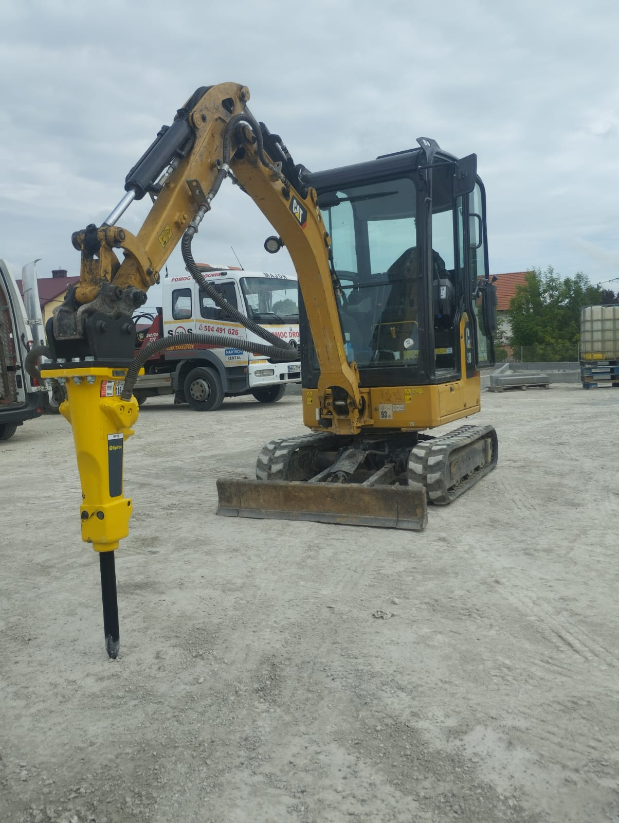 Żółta minikoparka CAT z zamontowanym młotem hydraulicznym Epiroc na placu budowy, w tle biała laweta z napisem Pomoc Drogowa.