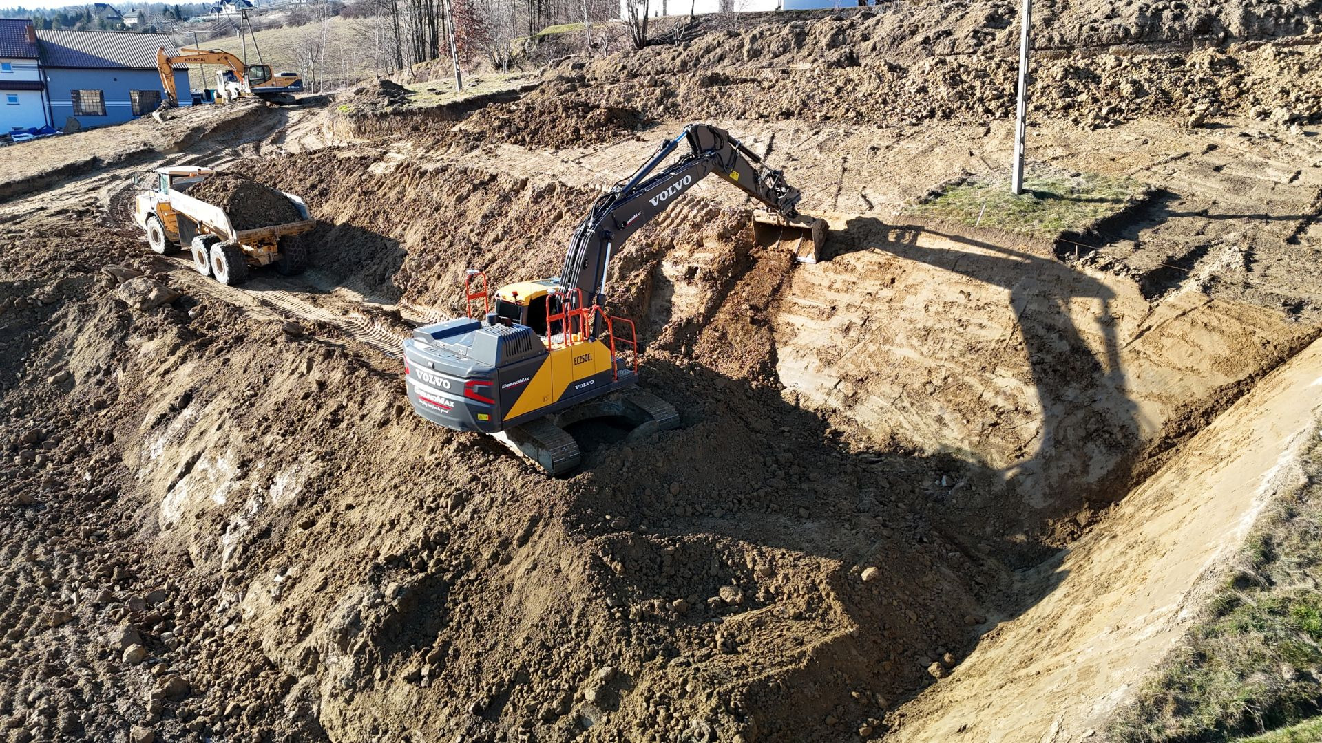 Żółto-czarna koparka Volvo EC250EL w trakcie prac ziemnych, wykop pod fundamenty, widoczna ciężarówka wywożąca urobek, w tle dom jednorodzinny i koparka Hyundai.