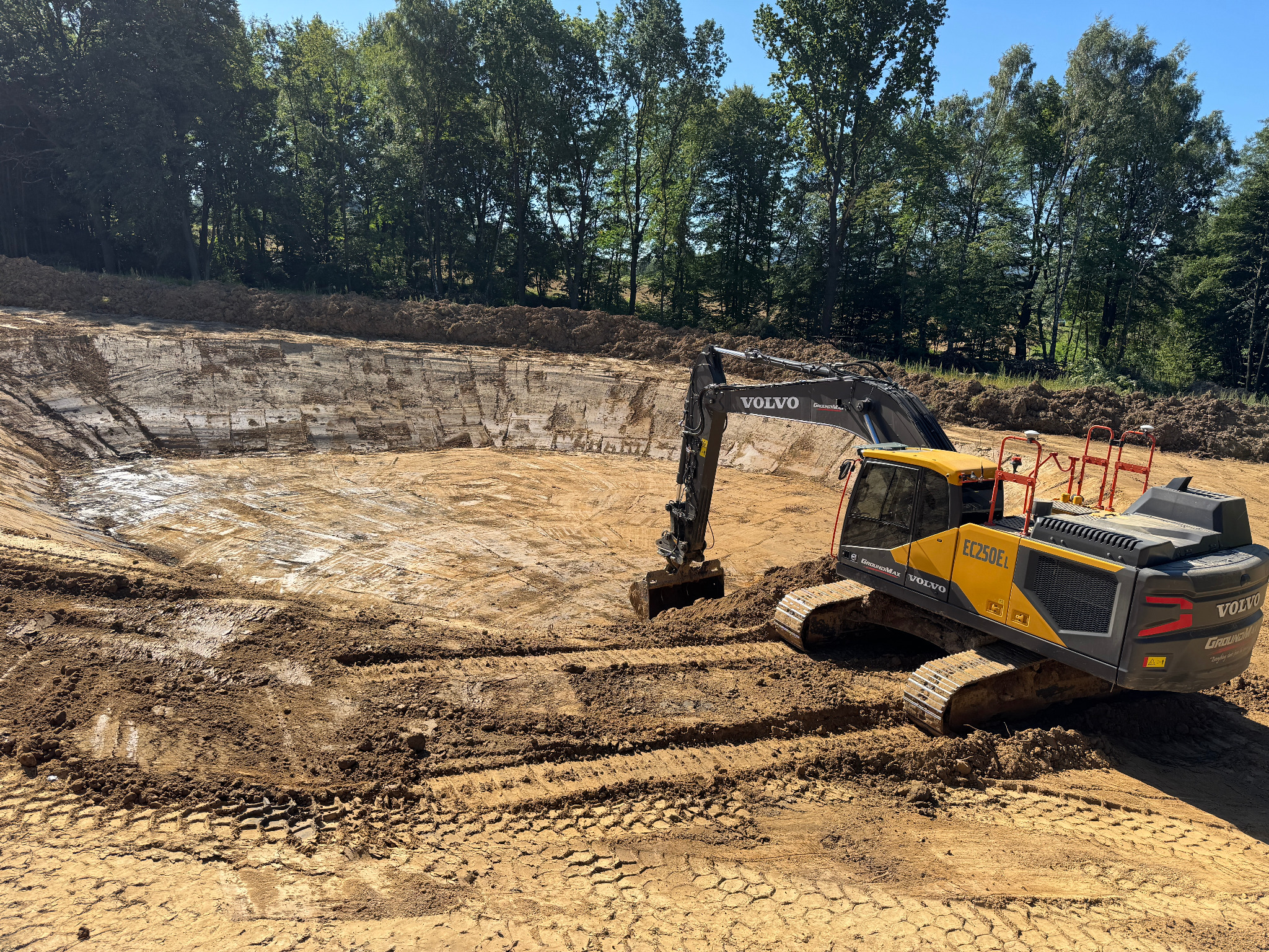 Żółta koparka Volvo EC250EL w wykopie, prawdopodobnie podczas wykopywania stawu. Ślady opon na ziemi, w tle drzewa i błękitne niebo.