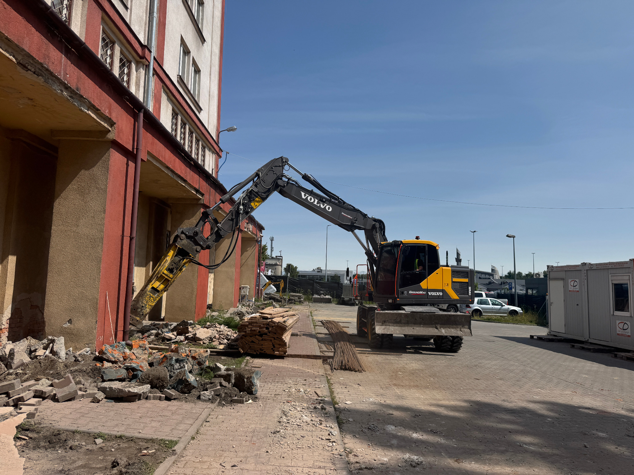 Żółto-czarna koparka Volvo z hydraulicznym młotem przy budynku w trakcie rozbiórki. Gruz i materiały budowlane na pierwszym planie, błękitne niebo w tle.