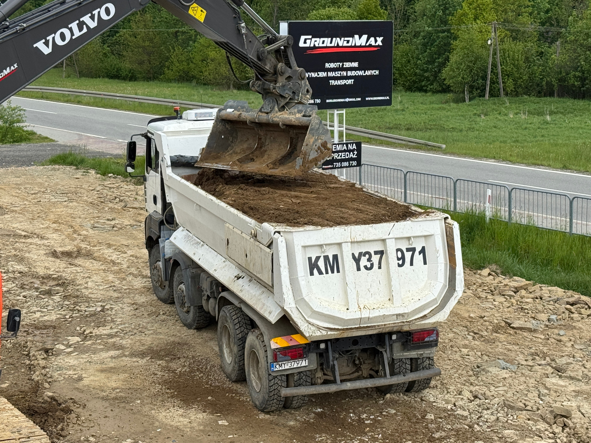 Koparka Volvo ładuje ziemię do wywrotki na tle billboardu GroundMAX, oferującej roboty ziemne i wynajem maszyn budowlanych. Widok z tyłu na ciężarówkę.