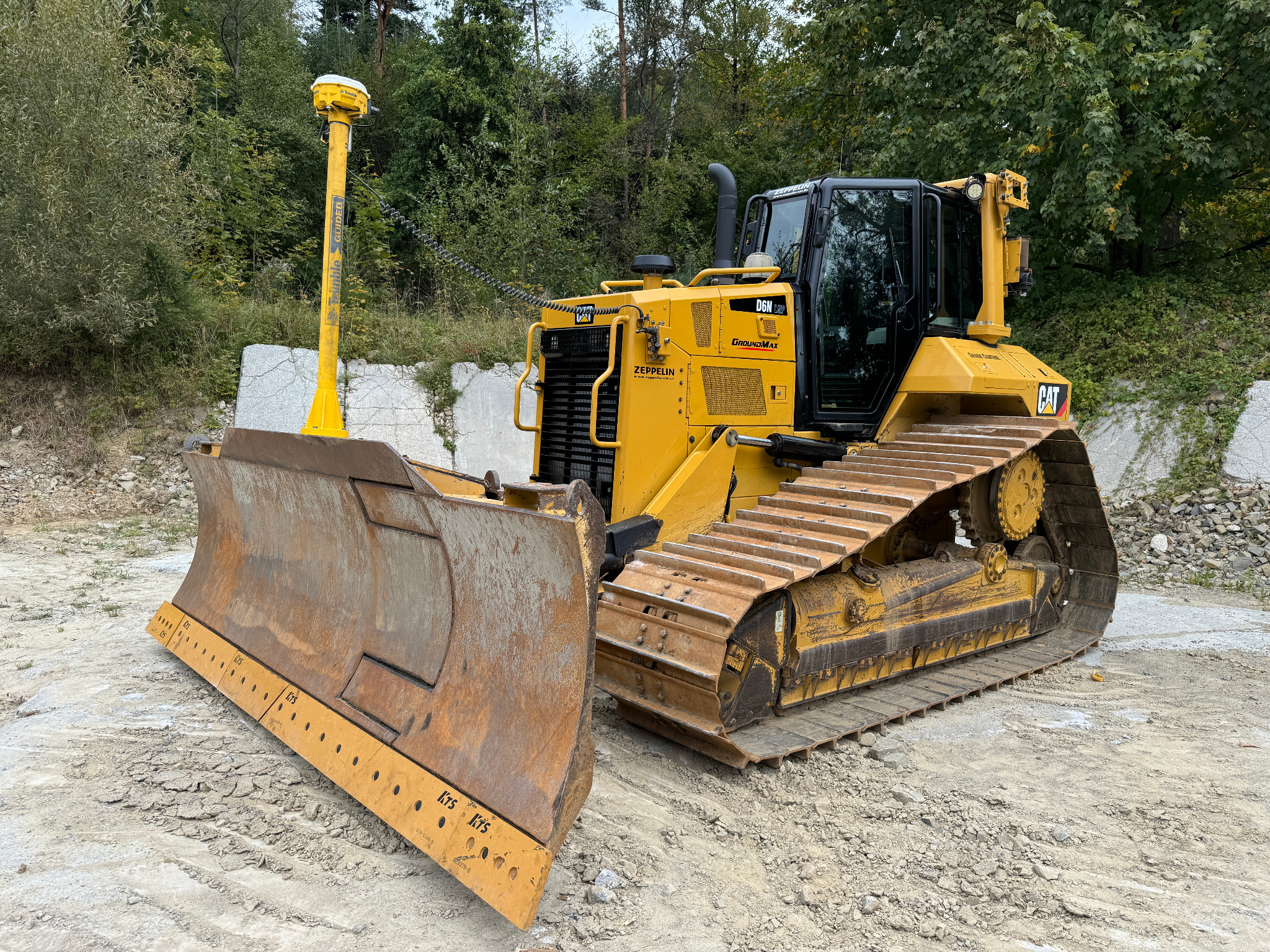 Żółty spychacz Caterpillar D6N LGP z systemem Trimble Guided na tle zieleni i betonowych bloków. Widoczne ślady pracy na ziemi.