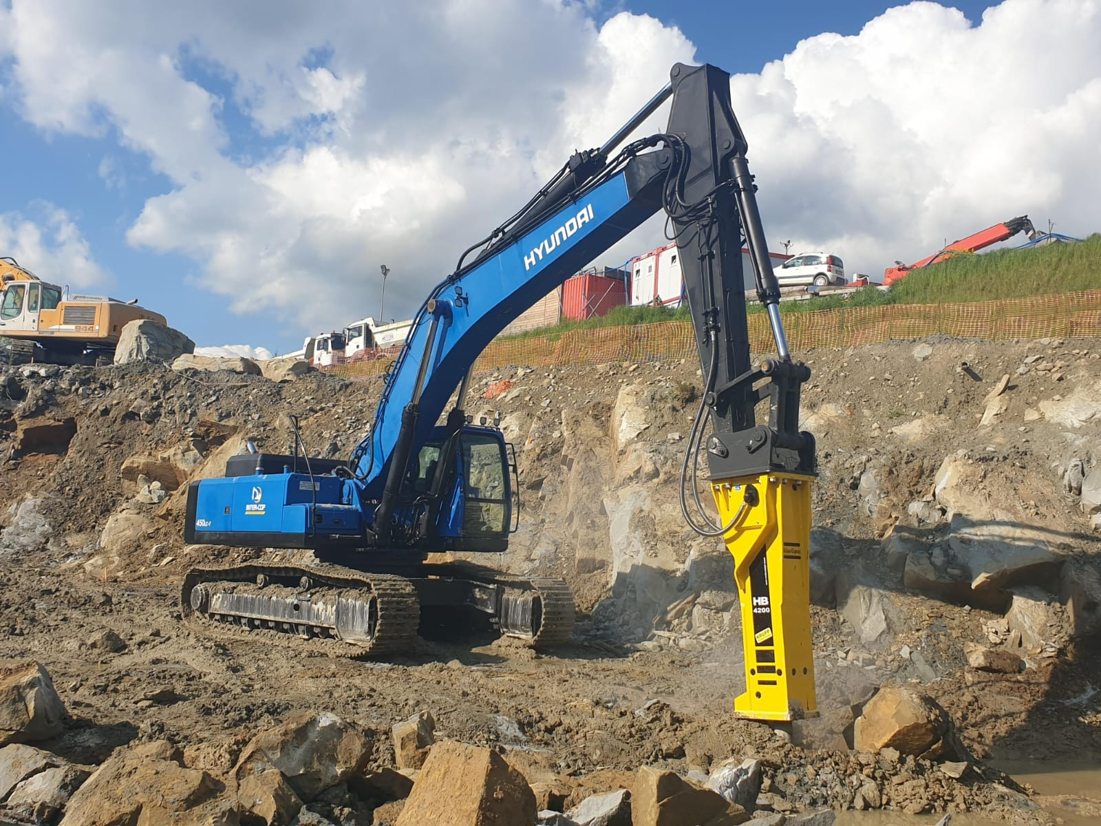 Niebieska koparka Hyundai z młotem hydraulicznym w akcji w Krzeczowie. Sprzęt na tle wykopu i błękitnego nieba z chmurami. Widoczne inne maszyny budowlane.