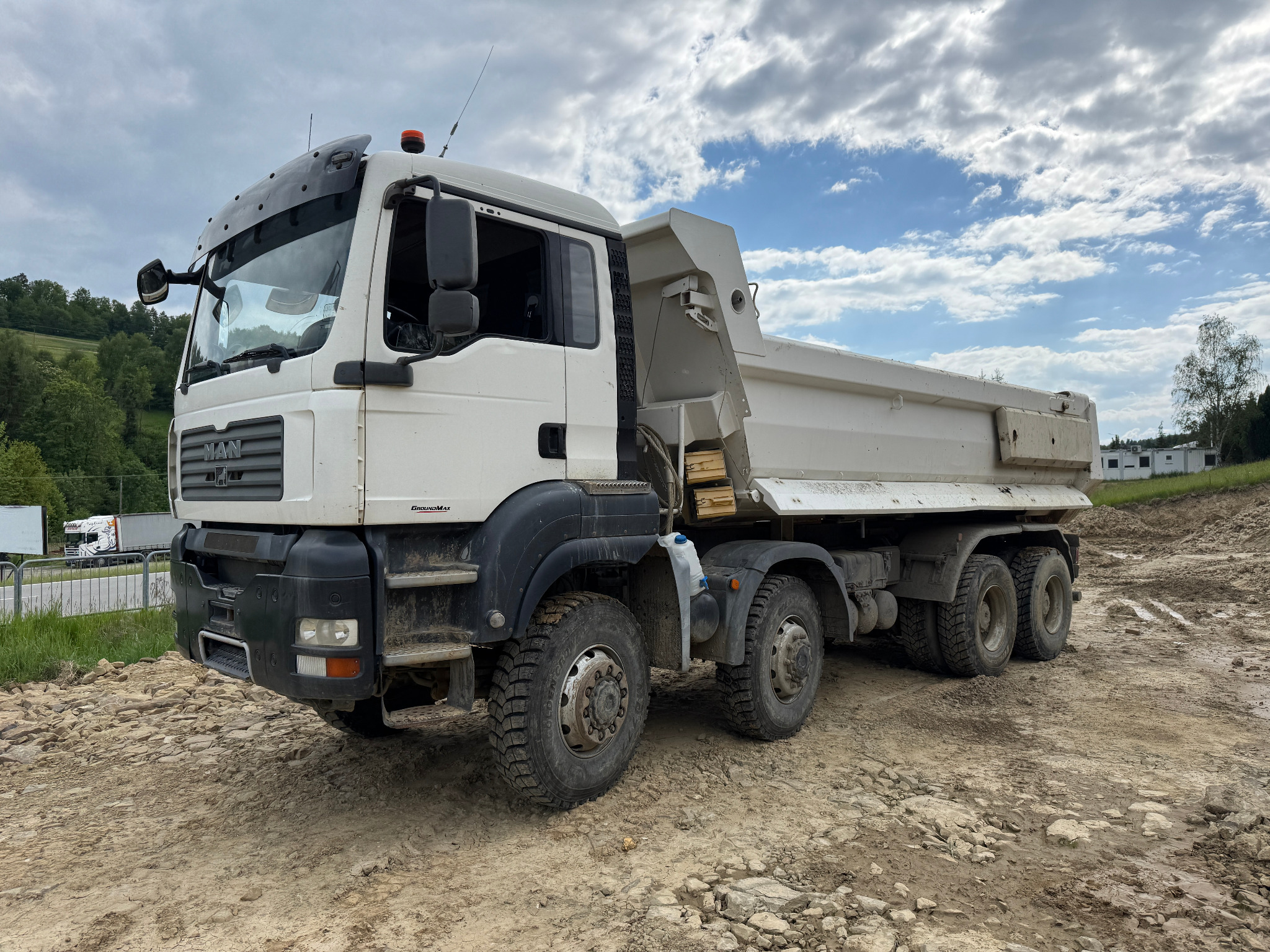 Biała ciężarówka MAN z wywrotką na nierównym, błotnistym terenie budowy, z widocznymi śladami opon i fragmentem zieleni w tle pod błękitnym niebem.