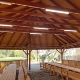 Zadaszona altana z drewnianymi ławami i stołami, oświetlona szeregiem lamp jarzeniowych pod drewnianym stropem, z widokiem na park na zewnątrz.