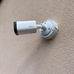 Biała kamera monitoringu Dahua zamontowana na beżowej, chropowatej ścianie budynku, widoczne śruby mocujące i logo producenta.