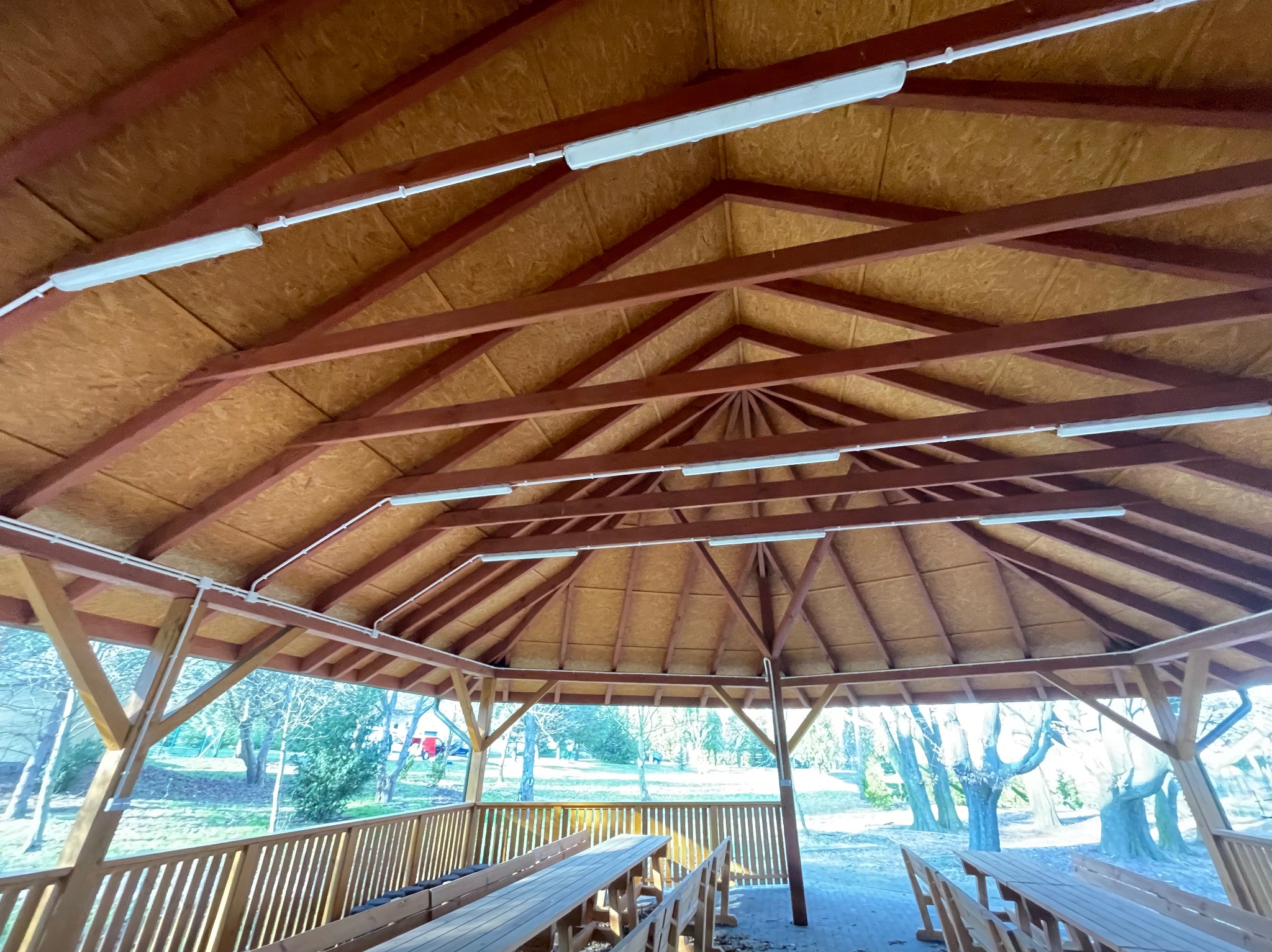 Wnętrze altany z drewnianą konstrukcją dachu i zamontowanymi lampami LED, widok na zewnątrz przez drewniane barierki.