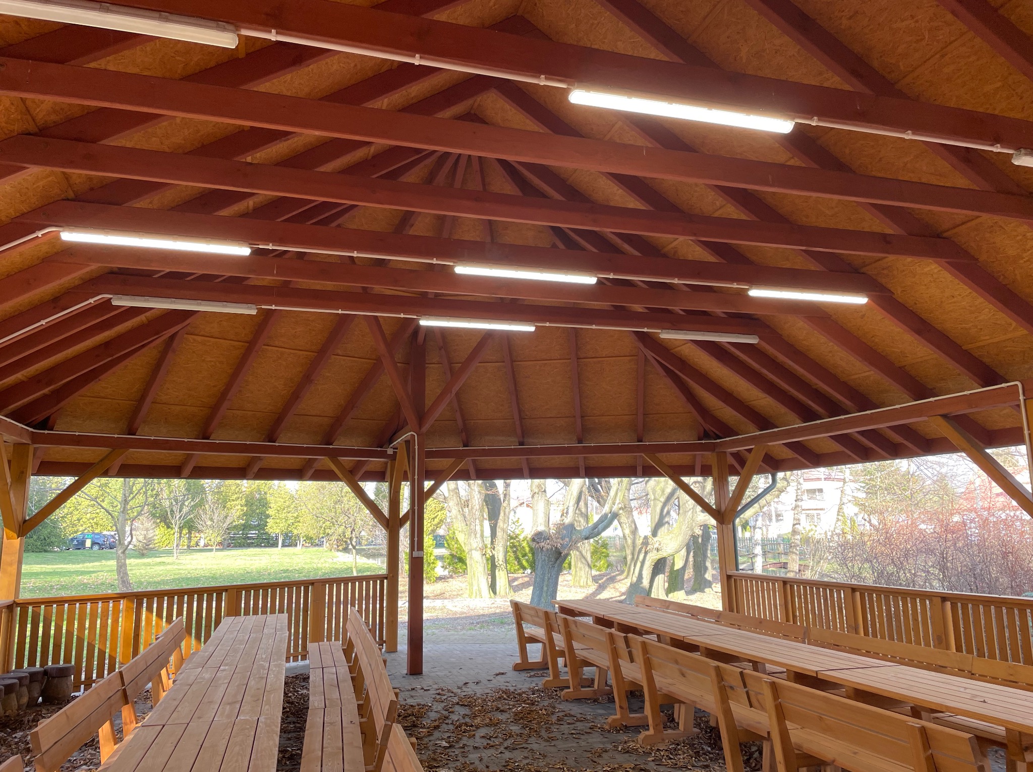 Zadaszona altana z drewnianymi ławami i stołami, oświetlona szeregiem lamp jarzeniowych pod drewnianym stropem, z widokiem na park na zewnątrz.