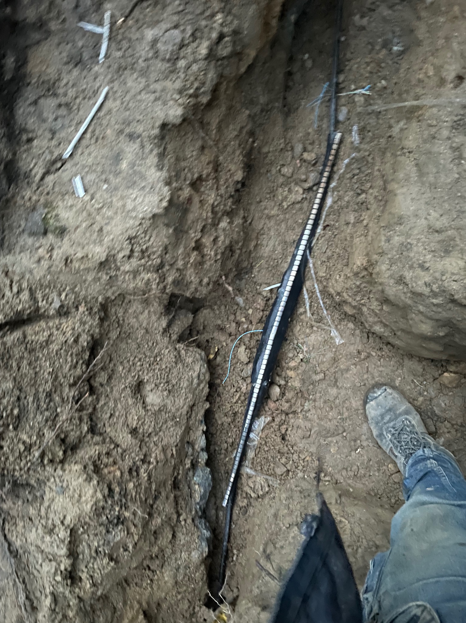 Kabel zakopany w wykopie ziemnym, widoczny fragment buta i spodni osoby instalującej.