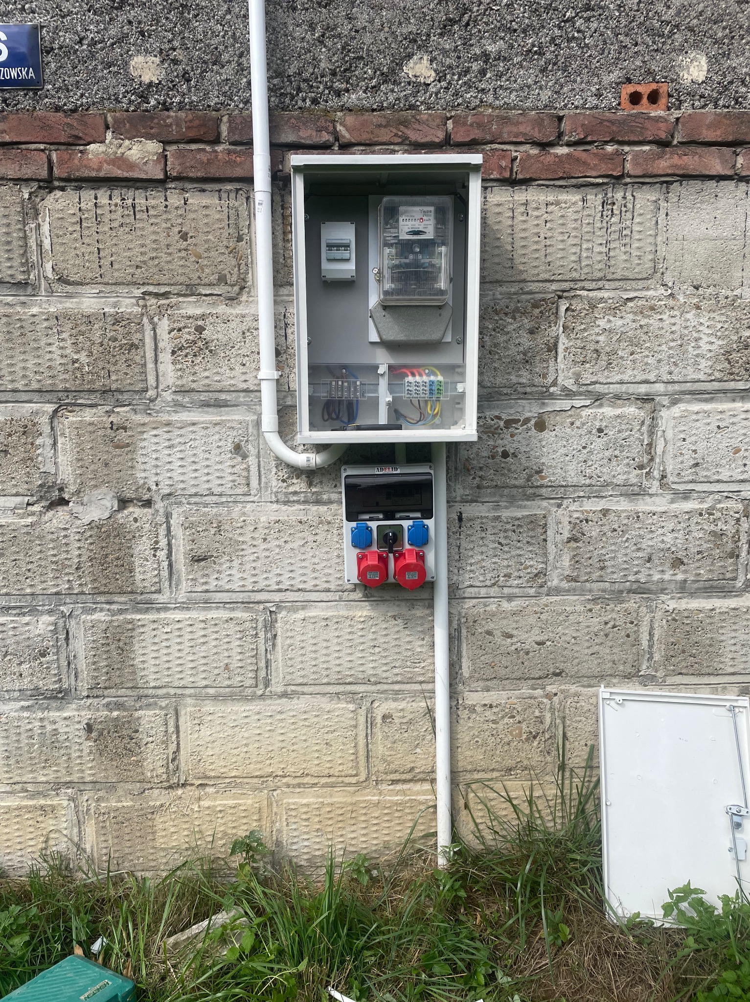 Zewnętrzna skrzynka elektryczna z licznikiem i gniazdami siłowymi, zamontowana na szarej, blokowej ścianie z ceglanym pasem, widoczne rury instalacyjne i trawa u podstawy.