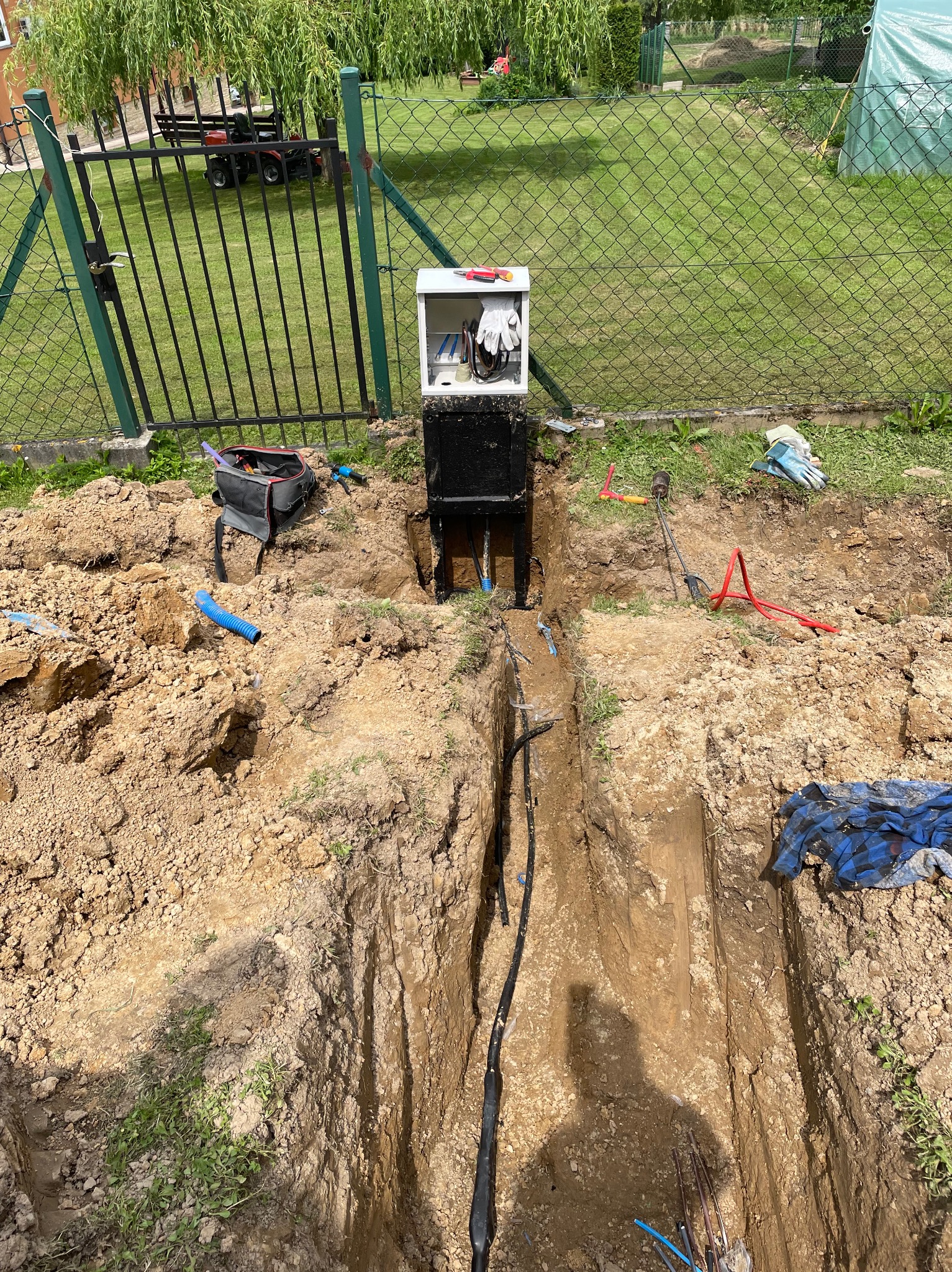 Wykop w ziemi z ułożonymi kablami elektrycznymi, skrzynka z narzędziami i rękawicami na słupku, teren posesji z ogrodzeniem w tle.