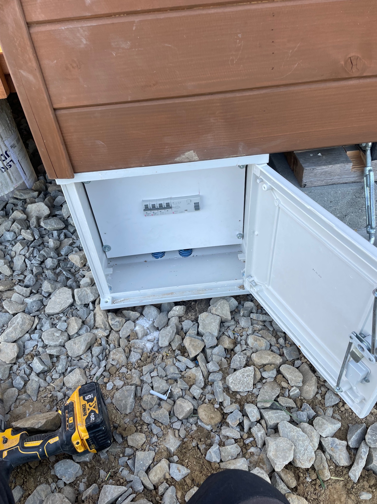 Otwarta skrzynka elektryczna z zamontowanymi bezpiecznikami, umieszczona pod drewnianą konstrukcją, widoczna w otoczeniu kamieni i narzędzia - żółto-czarna wiertarka akumulatorowa.