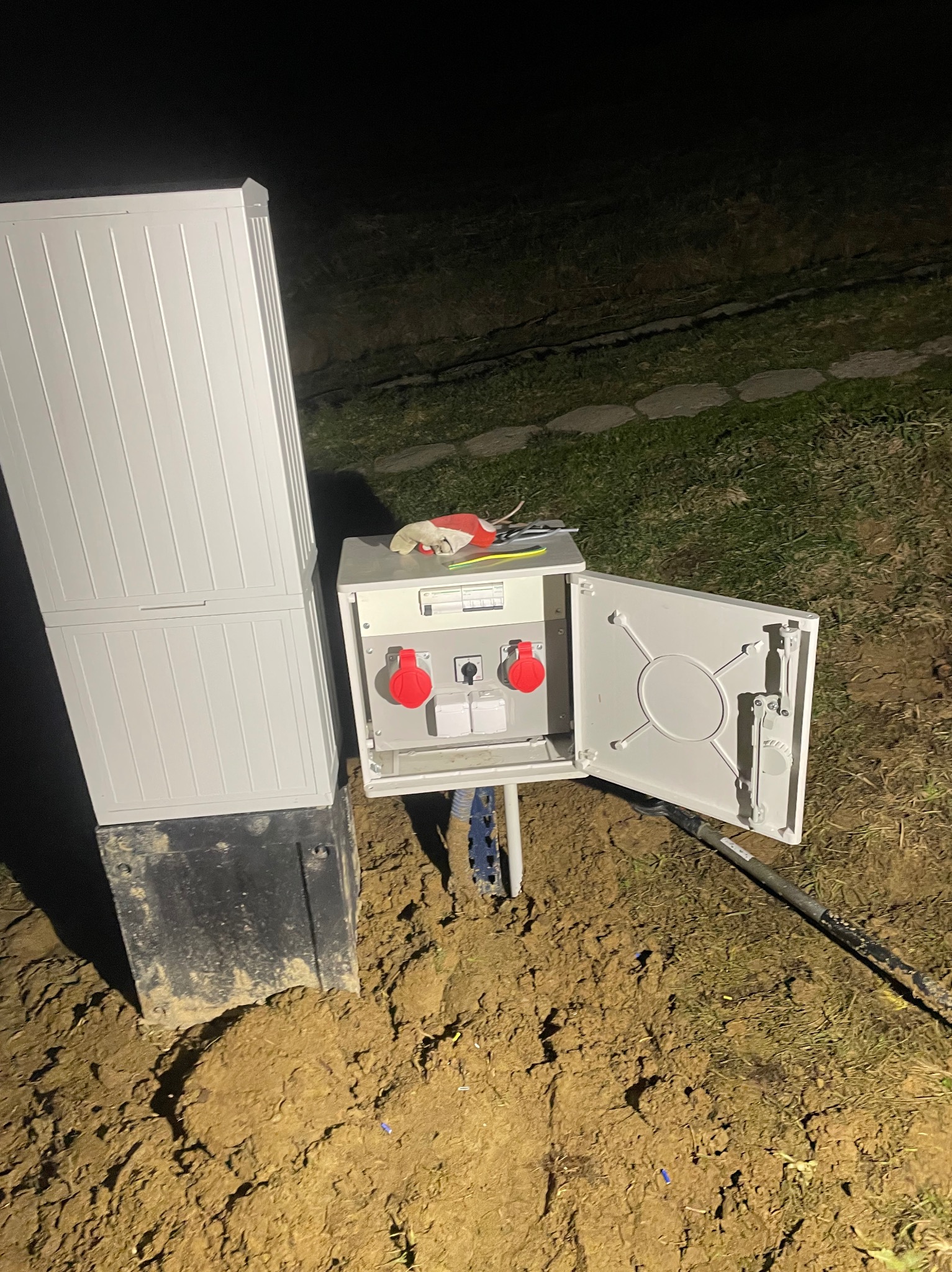 Zewnętrzna skrzynka elektryczna z otwartymi drzwiczkami, zamontowana na fundamencie w ziemi, widoczne gniazda siłowe i zabezpieczenia, obok narzędzia i rękawice robocze.