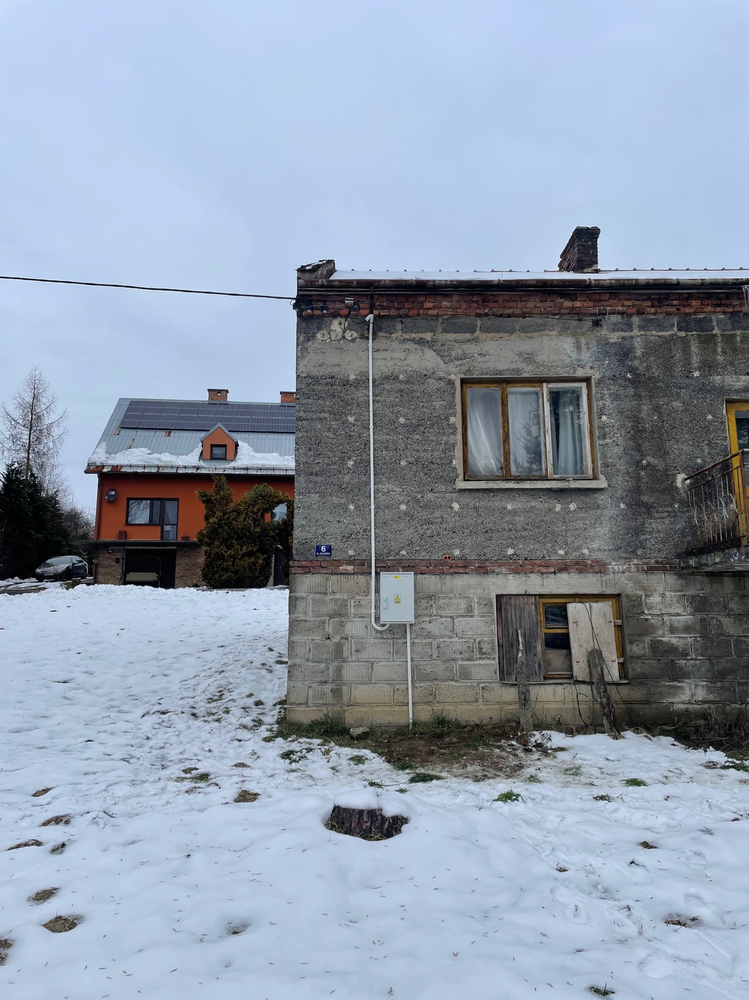 Szary budynek z widoczną skrzynką elektryczną na zewnątrz, na tle ośnieżonego terenu i budynku z panelami słonecznymi na dachu.