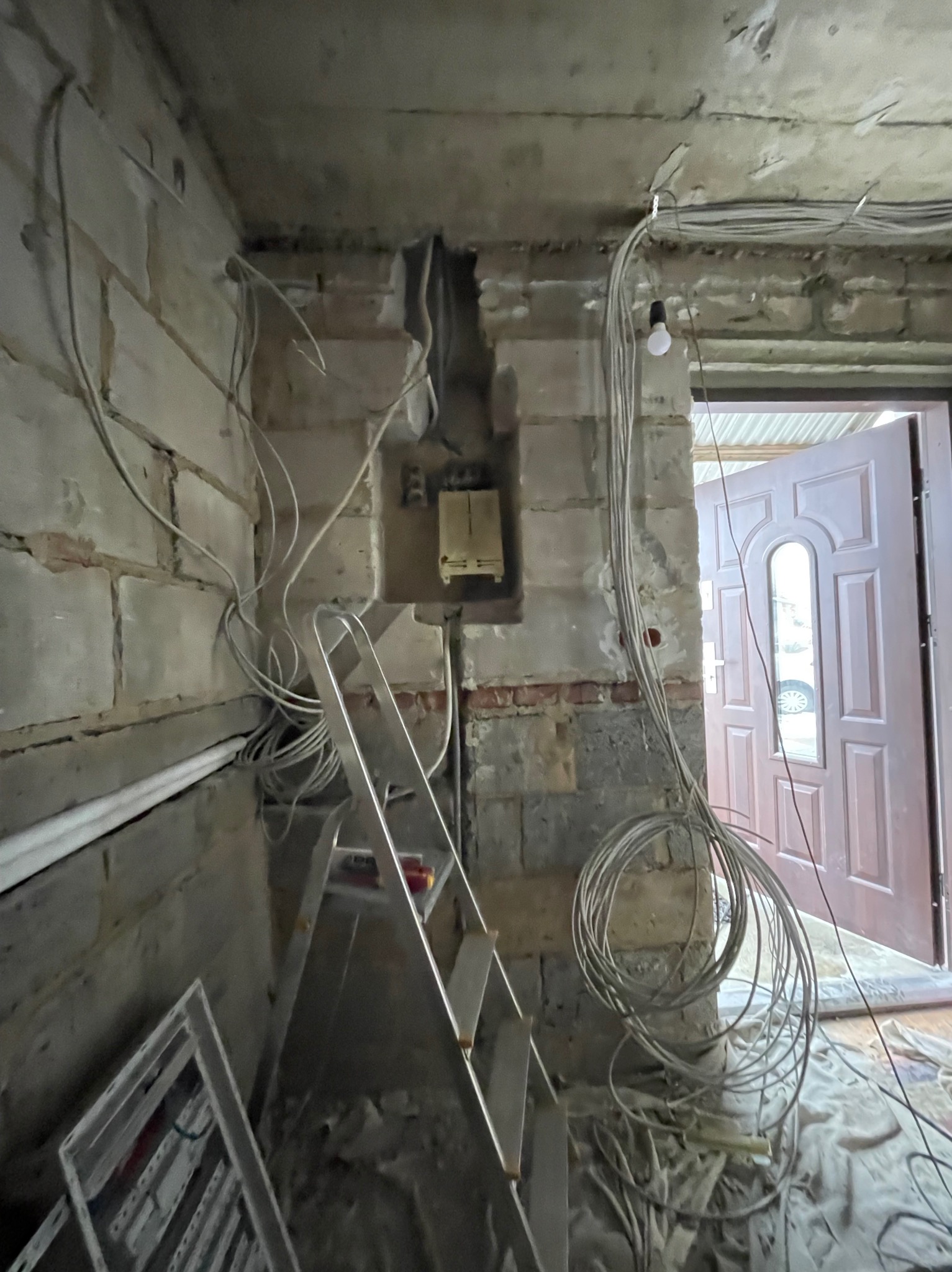 Surowy stan instalacji elektrycznej w budynku z odsłoniętą cegłą, widoczne przewody, skrzynka elektryczna w ścianie i drabina aluminiowa.