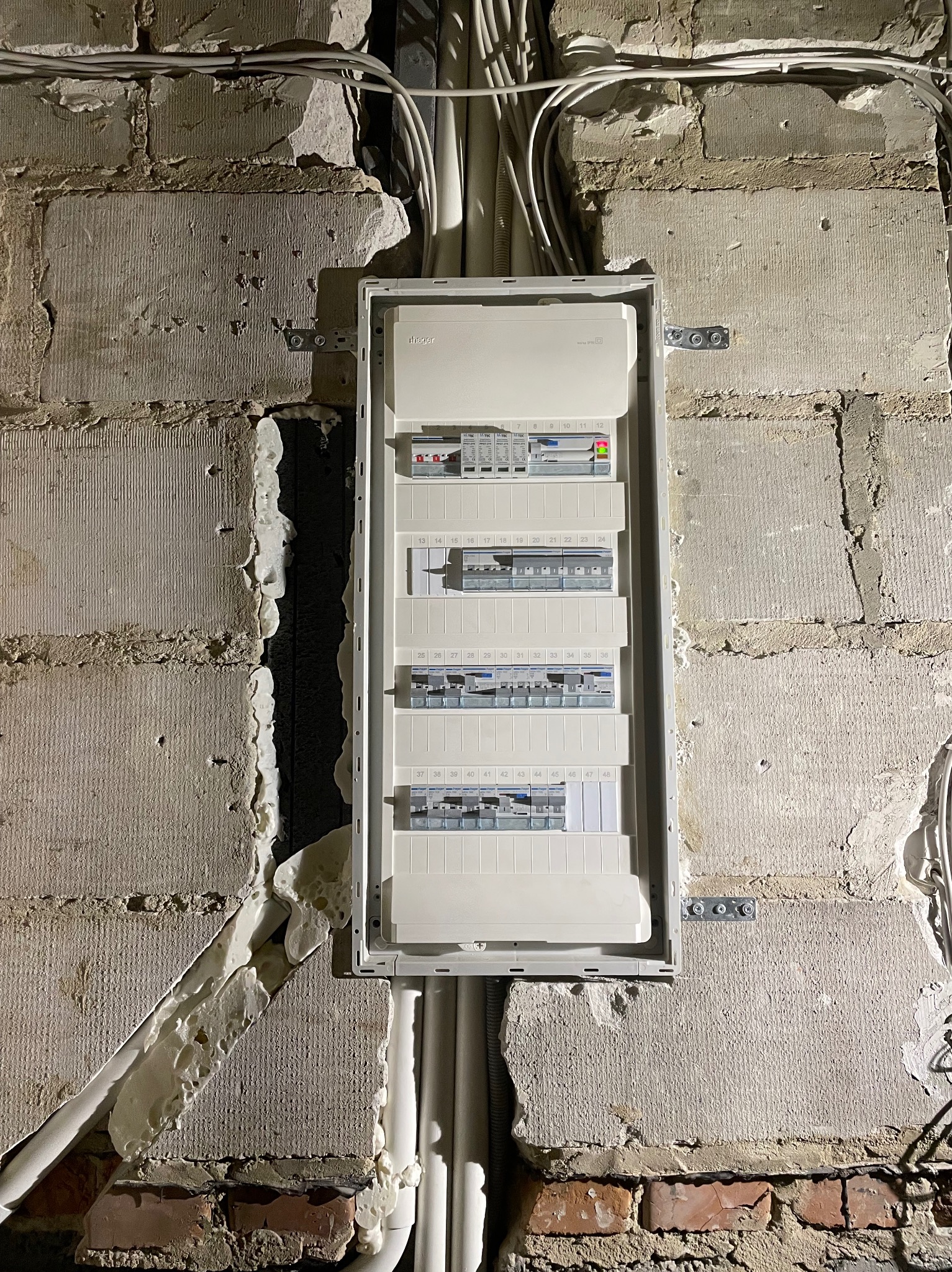 Rozdzielnica elektryczna zamontowana na surowej ścianie z betonowych pustaków i cegieł, z widoczną pianką montażową wokół, oraz prowadzącymi do niej kablami w peszlach.